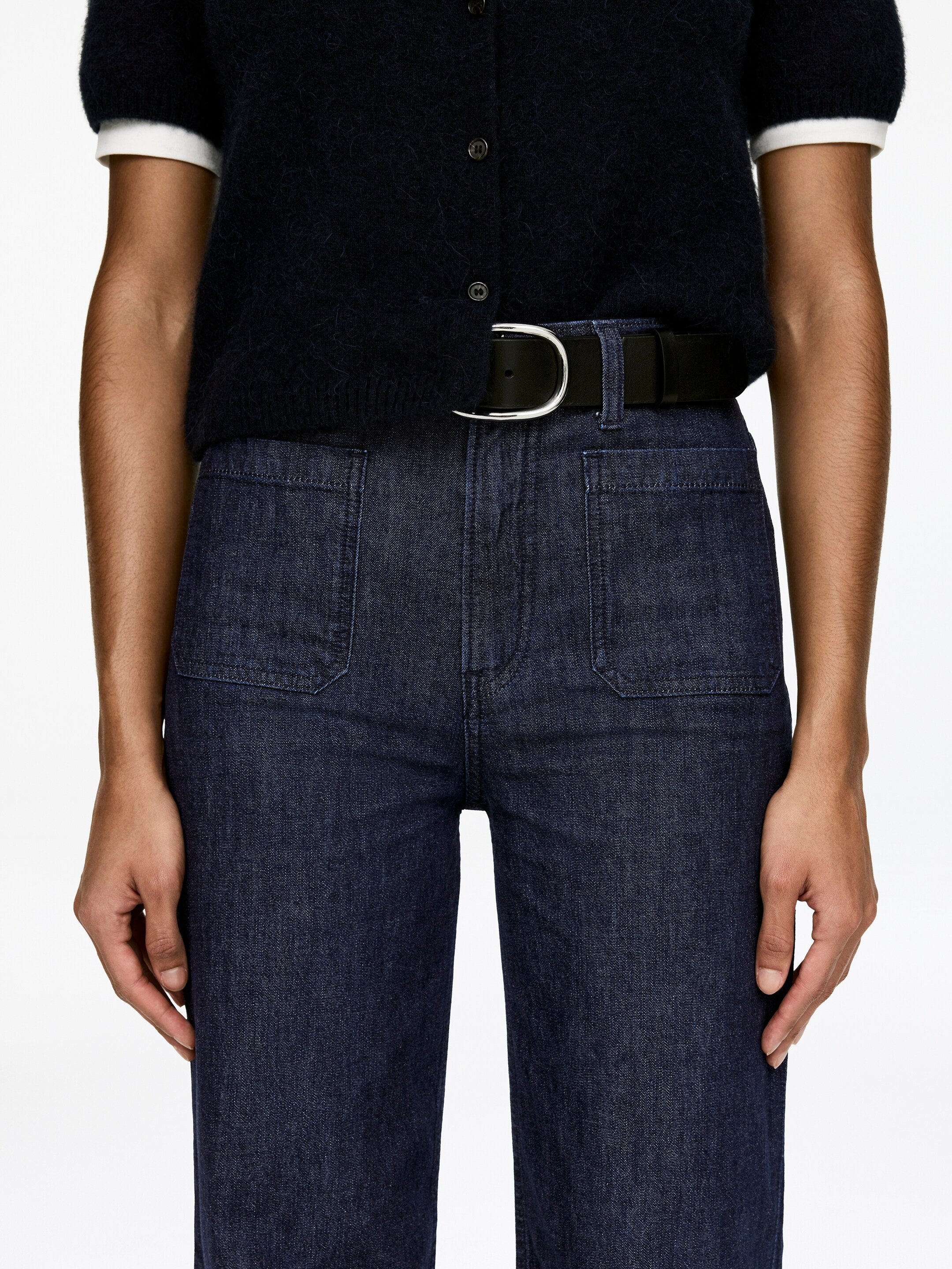 Agrandir l'image: Des jeans en denim indigo foncé sont présentés de face, avec une taille haute dotée de passants de ceinture et deux grandes poches plaquées sur le devant des cuisses, soulignées par une couture contrastante. Une ceinture noire avec une boucle en D en argent est passée à travers les passants, cintrant la taille.