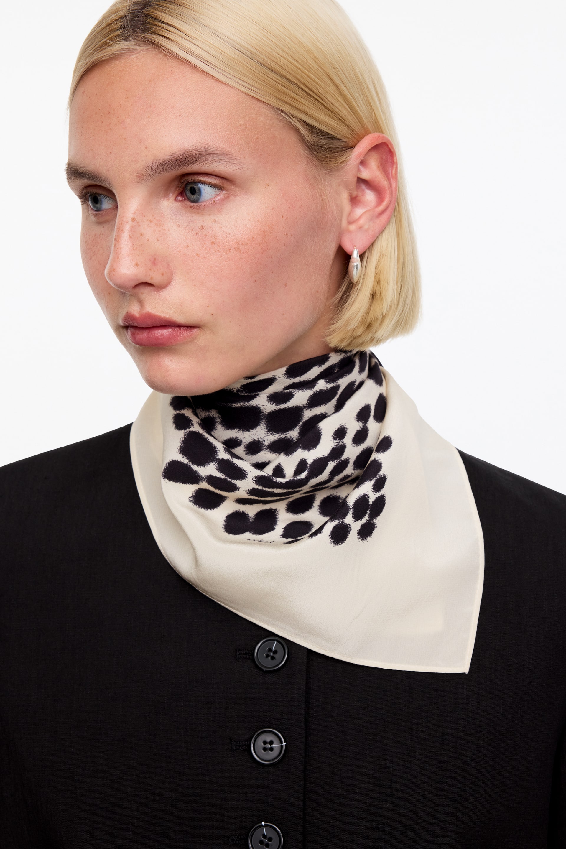 Grotere afbeelding bekijken: Een zachte, crèmekleurige sjaal met een onregelmatig zwart vlekkenpatroon hangt om de nek en rust over een zwart kledingstuk.
