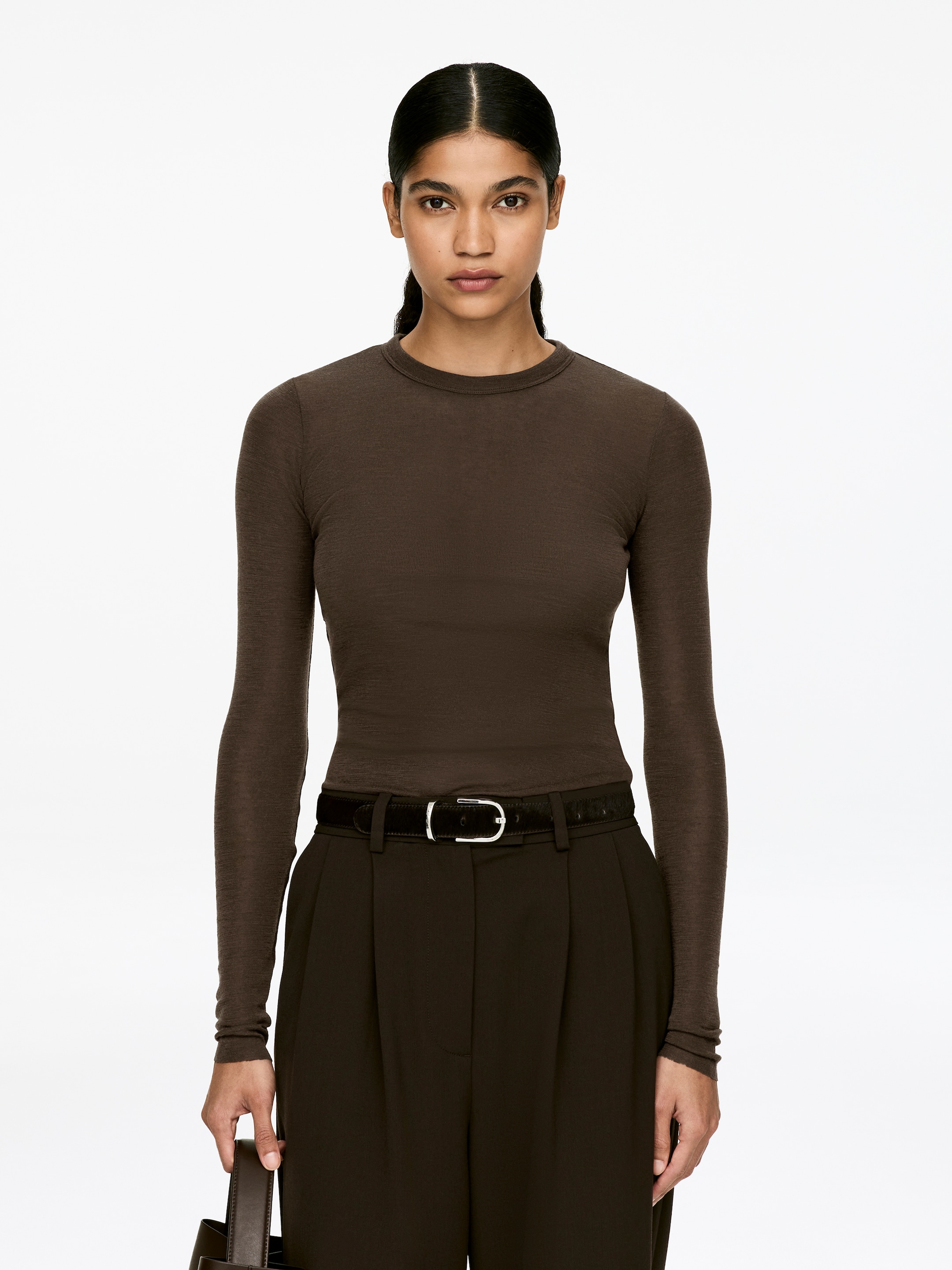 View larger image: A dark brown, tight-fitted merino wool top with a crew neck and long sleeves is being worn by the model. It has a slightly transparent appearance and features raw-cut sleeve ends, styled with dark brown pleated trousers and a black belt.