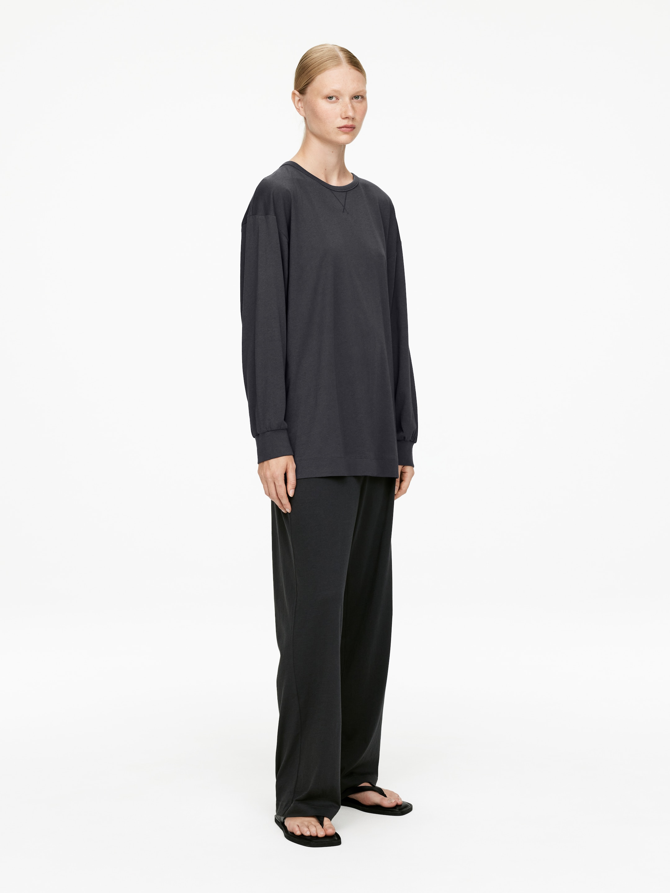 Agrandir l'image: Une chemise gris foncé à manches longues et un pantalon large assorti sont présentés sur un mannequin, légèrement incliné sur le côté. La chemise présente un col rond avec des surpiqûres visibles et des poignets élastiqués, complétant la coupe ample et large du pantalon.