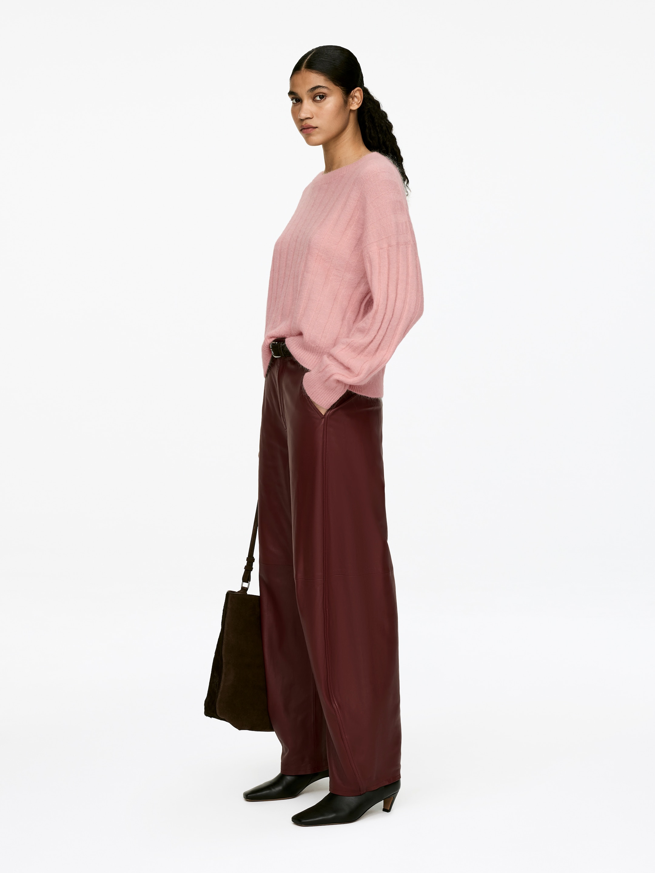 Grösseres Bild ansehen: Ein rosa Jumper mit breiten Rippen, der eine lässige Passform und einen Rundhalsausschnitt aufweist, wird von dem Model getragen. Das weich strukturierte Kleidungsstück ist teilweise in bordeauxrote Hosen gesteckt, wobei eine der Hände des Models in einer Hosentasche ruht.