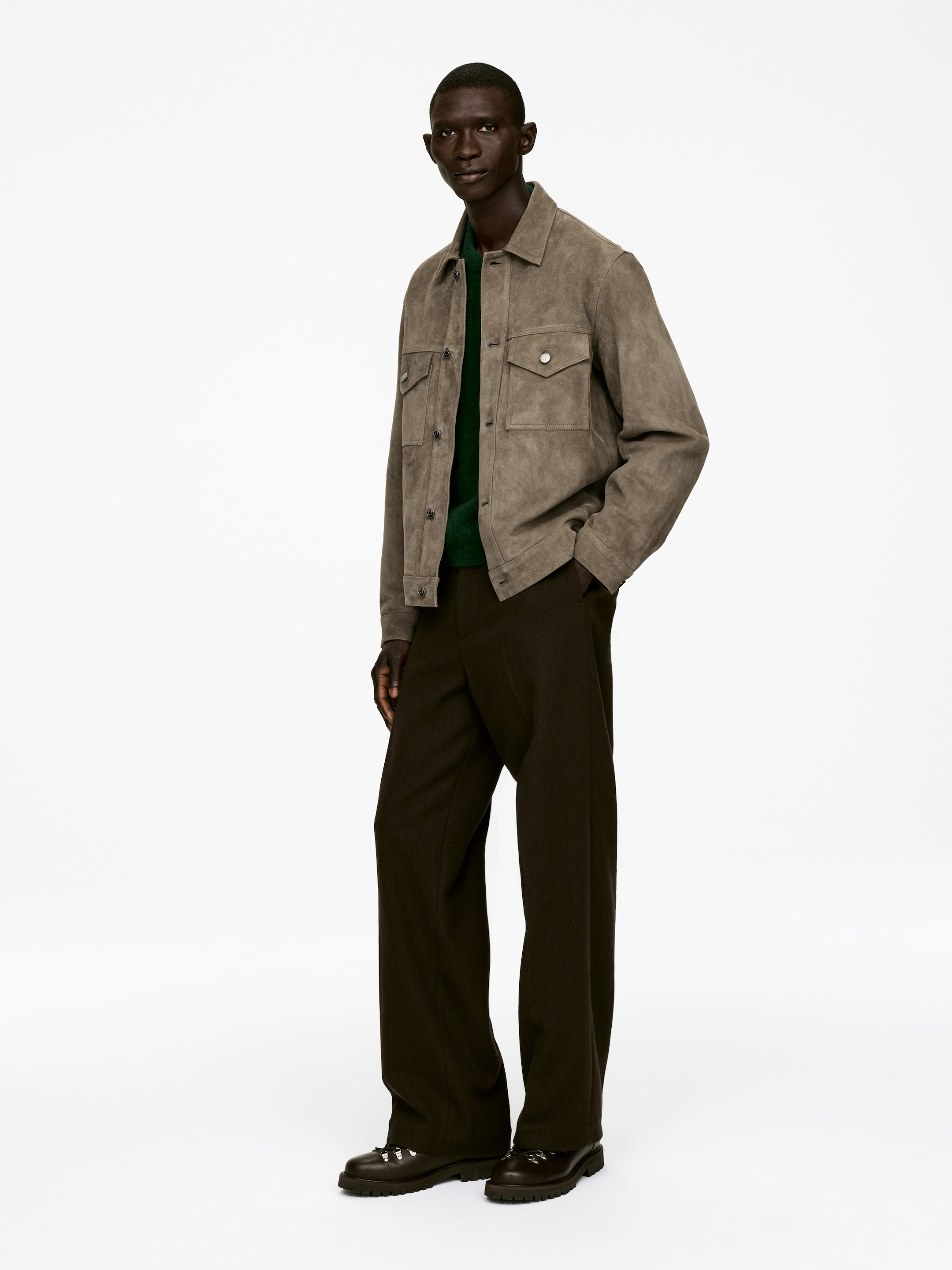 Agrandir l'image: Une veste-chemise en daim taupe présente un col pointu, deux poches poitrine à rabat boutonné et des poignets boutonnés. Elle est portée ouverte sur un pull à col rond vert et est stylée avec un pantalon large marron foncé et des chaussures marron à lacets qui mettent en valeur des semelles à crampons proéminentes et des anneaux en D argentés.