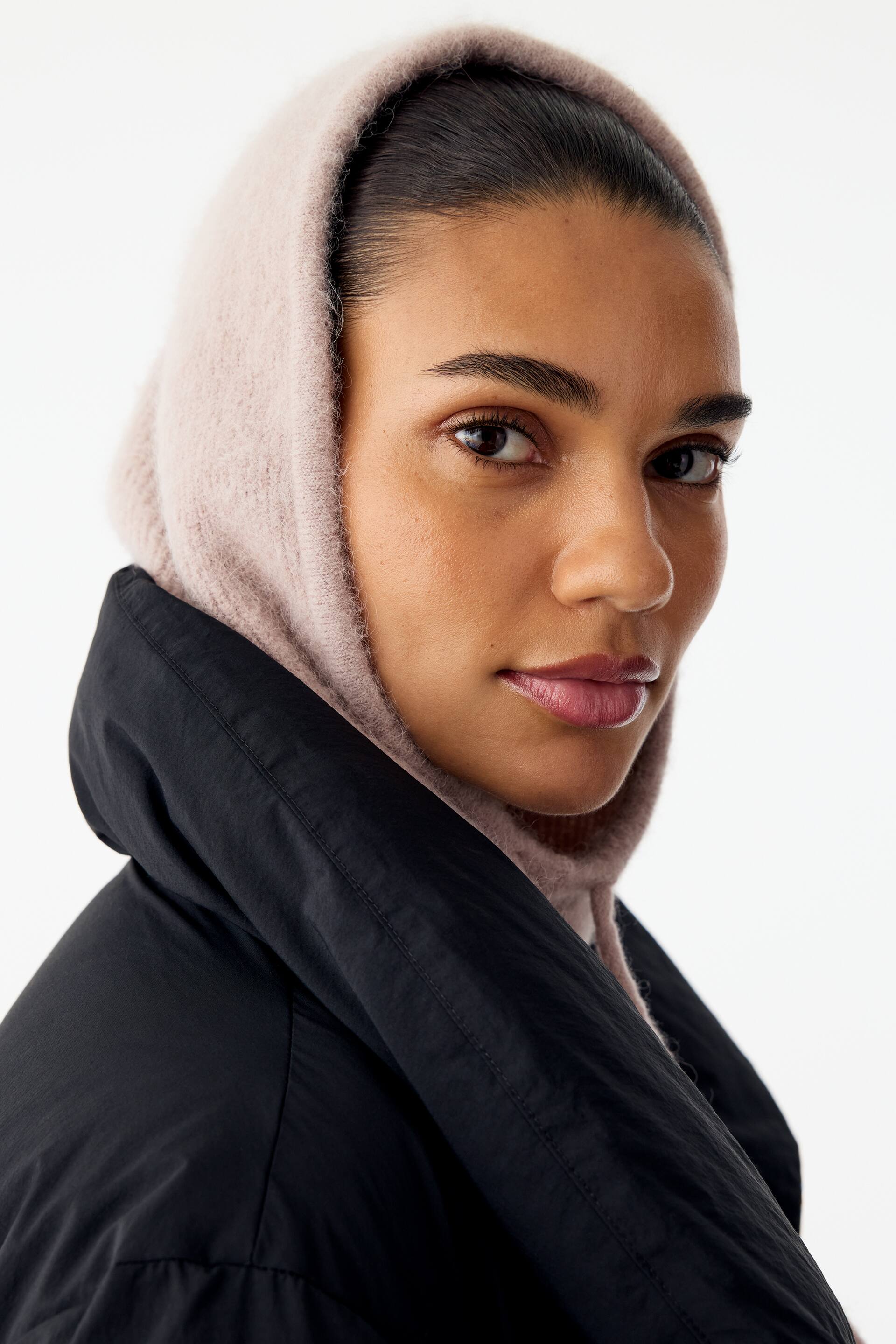 Agrandir l'image: Un capuchon en maille texturée rose clair est porté par le mannequin, encadrant son visage et son cou. Le capuchon est porté sous une veste noire dotée d’un col montant et matelassé.