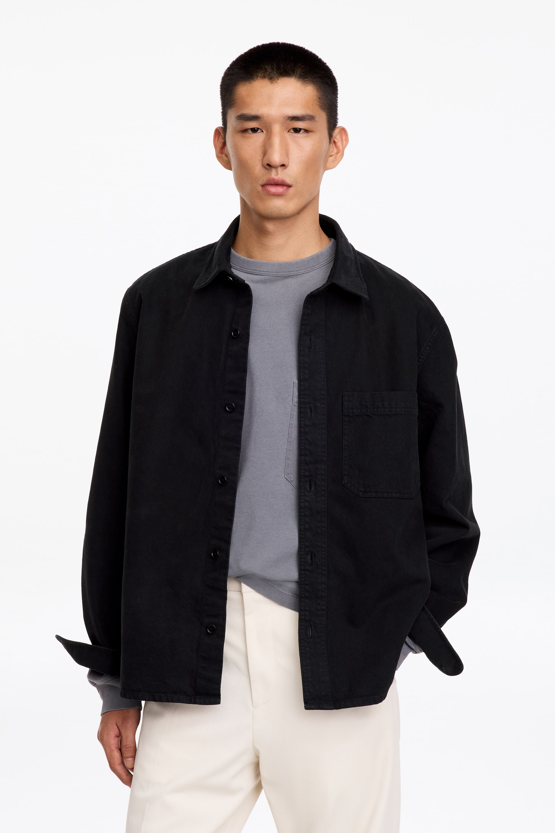 Visualizza l'immagine ingrandita: Una overshirt in twill nera è indossata aperta dalla modella, con colletto classico, una tasca applicata sul petto e polsini con bottoni. L’overshirt è abbinata a una t-shirt grigia a maniche lunghe e a pantaloni color bianco sporco.