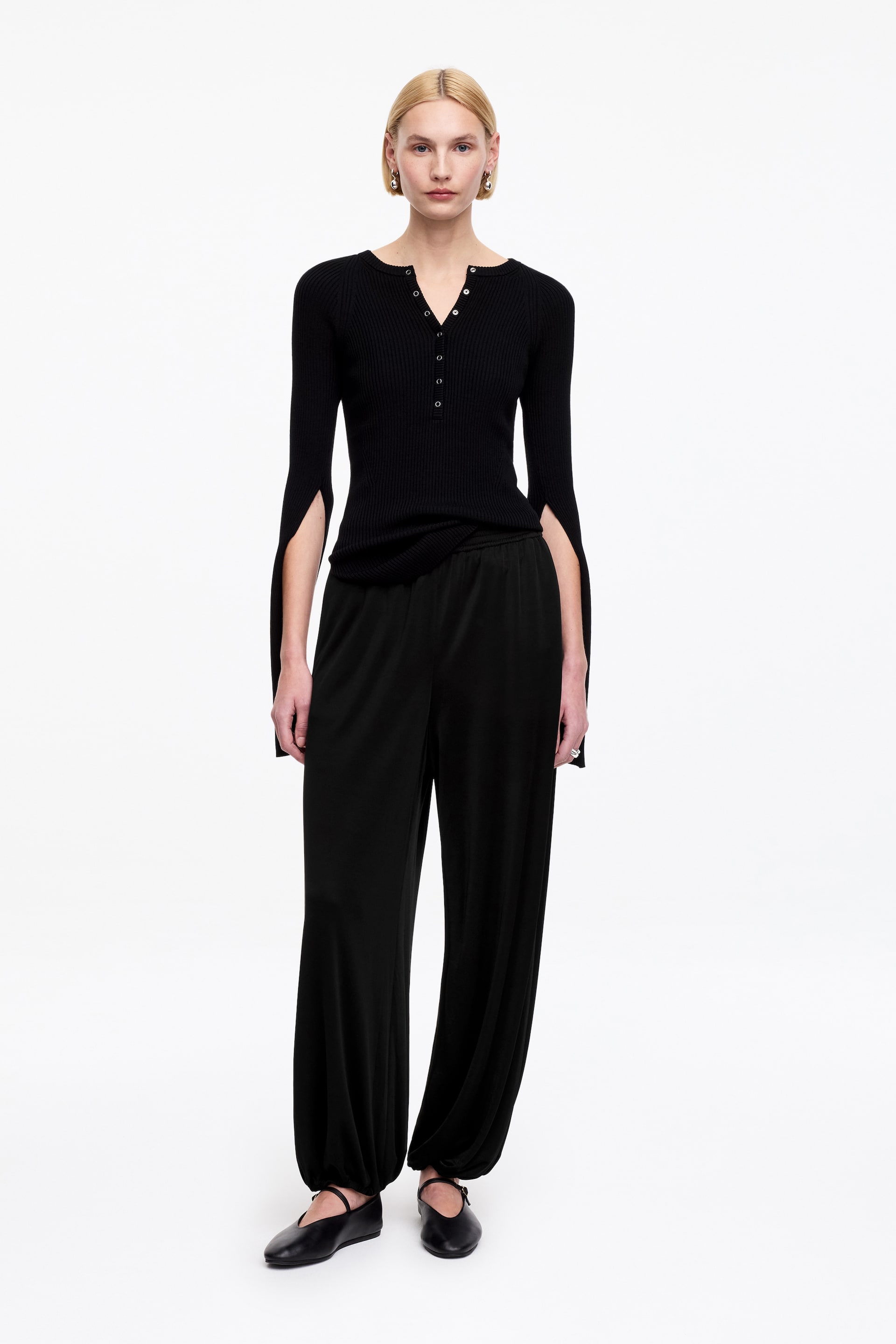 Nagyobb kép megtekintése: Egy fekete bordázott kötött henley felső V-nyakú gombolópánttal, több gombbal, hosszú ujjakkal, melyek külső alkarján hasítékok találhatók, teljes elölnézetben látható, fekete, laza szabású nadrággal, amely a bokánál rugalmas mandzsettával van összefogva.