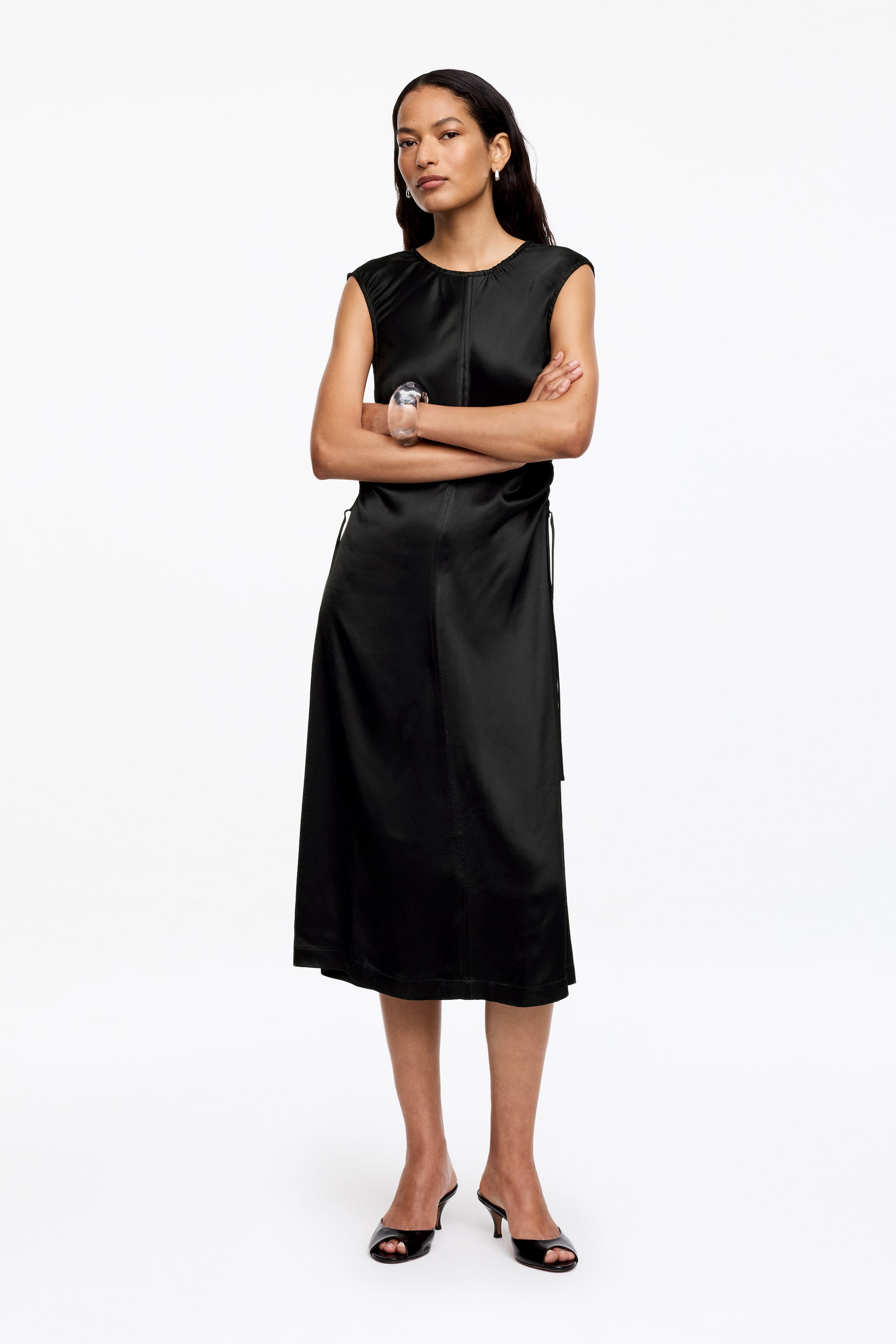 Visa större bild: En svart ärmlös midiklänning presenteras framifrån, med en rund halsringning och en glansig finish. Den är designad med en synlig vertikal söm längs mitten fram och en knytdetalj på vänster sida, vilket möjliggör en justerbar silhuett.