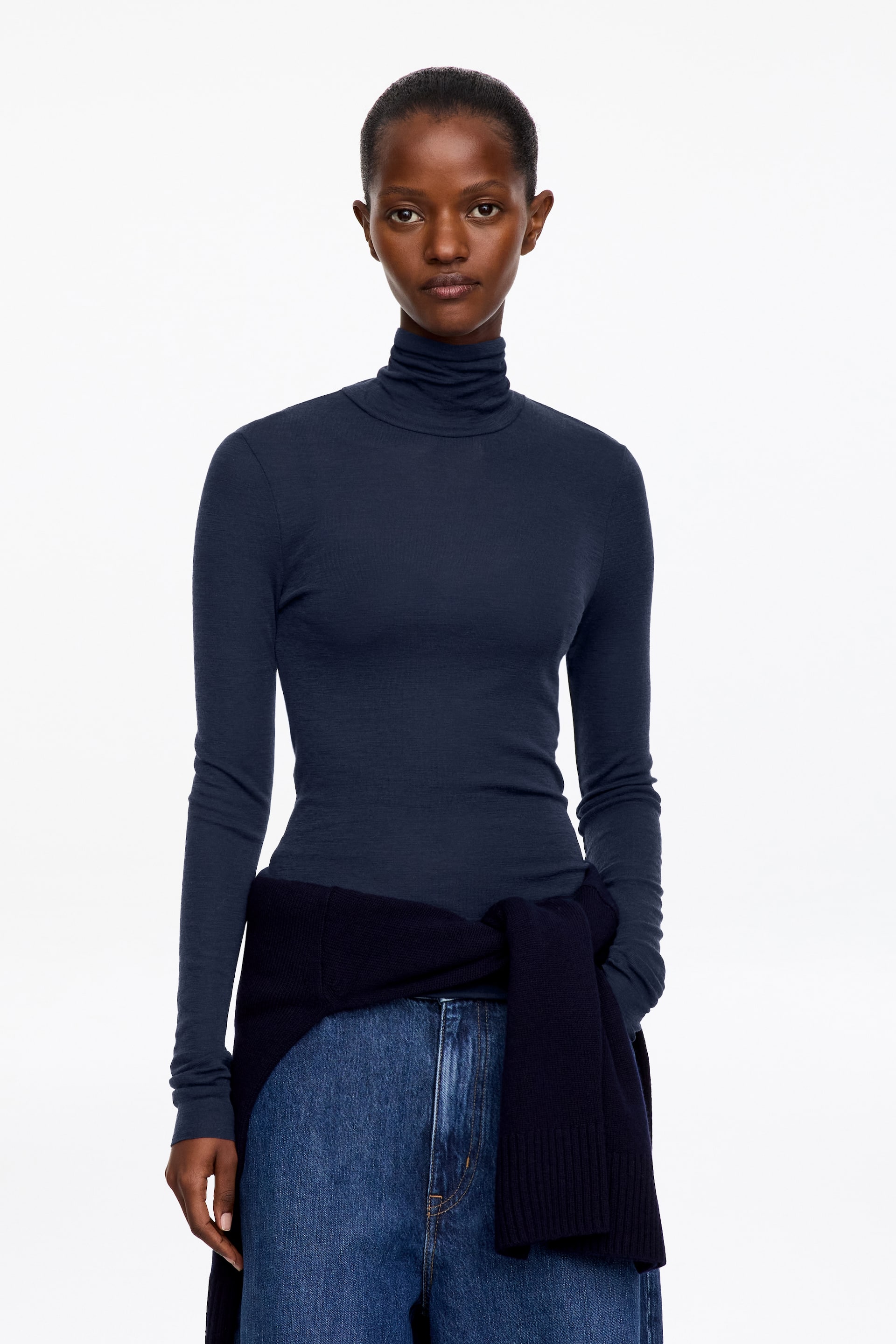 Vezi imaginea mai mare: Un top jersey cu guler înalt, transparent, de culoare albastru marin închis, este purtat de model, având o croială slim fit și mâneci lungi cu manșete cu margine netivită. Topul este stilizat cu blugi albaștri și un pulover tricotat închis la culoare, legat în jurul taliei.