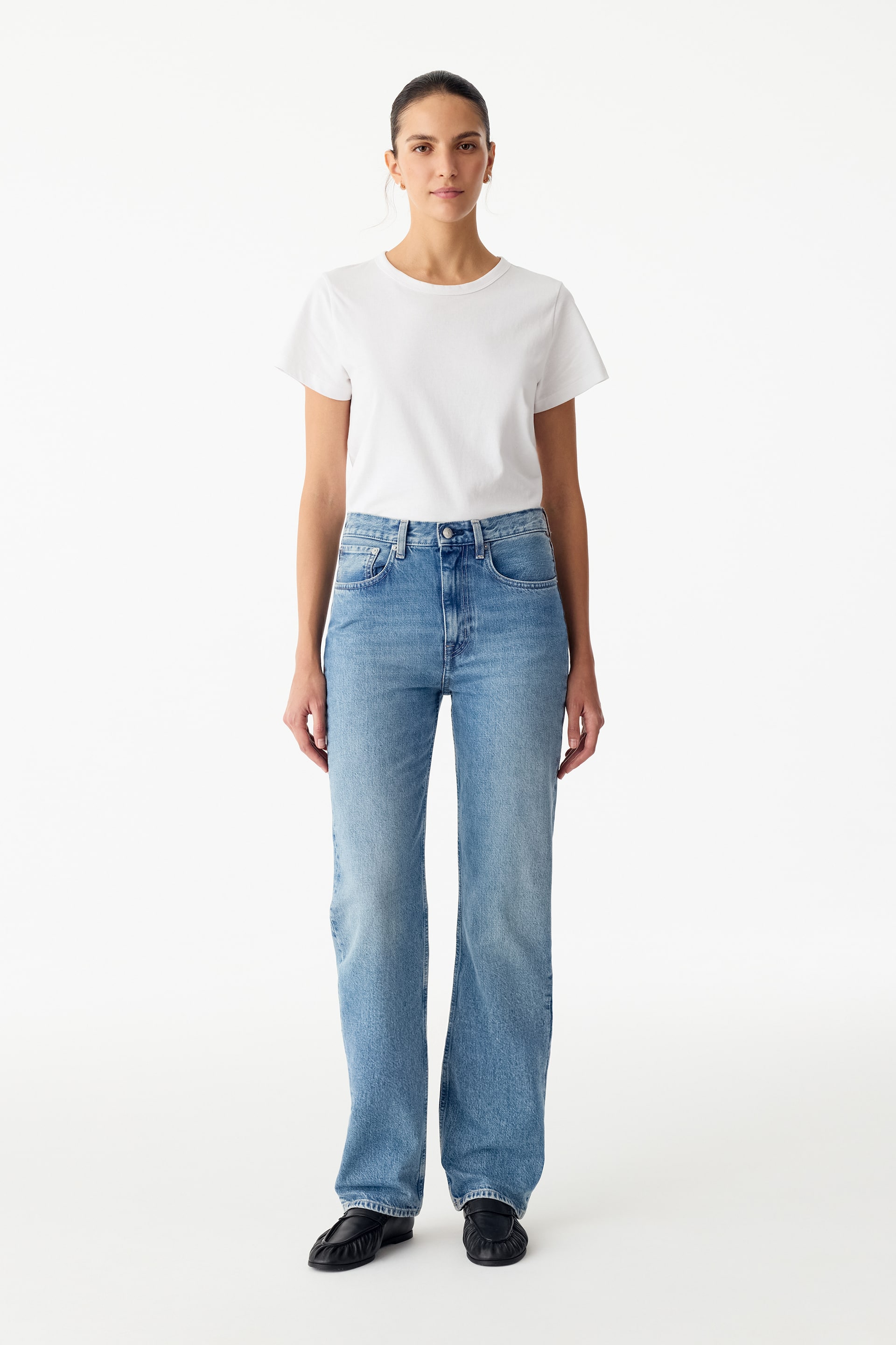 Agrandir l'image: Ce jean en denim bleu clair présente une taille mi-haute et une coupe droite, et il est porté par le mannequin. Le jean est associé à un t-shirt blanc à col rond rentré à l’intérieur.
