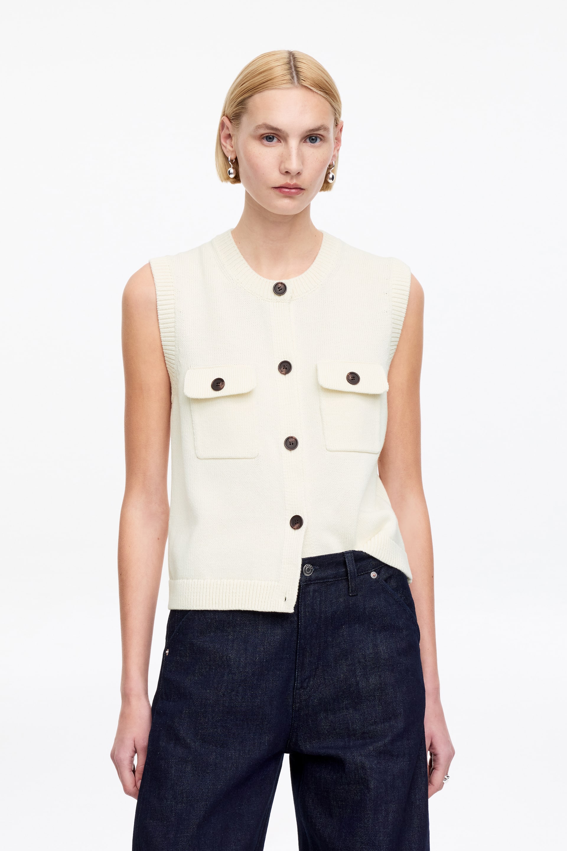 Visualizza immagine più grande: Il cardigan lavorato a maglia senza maniche color crema presenta uno scollo rotondo e una chiusura frontale completa con cinque bottoni marrone scuro. È progettato con due tasche applicate sul petto con patte, giromanica a coste e un orlo asimmetrico a coste parzialmente infilato nei jeans blu scuro.