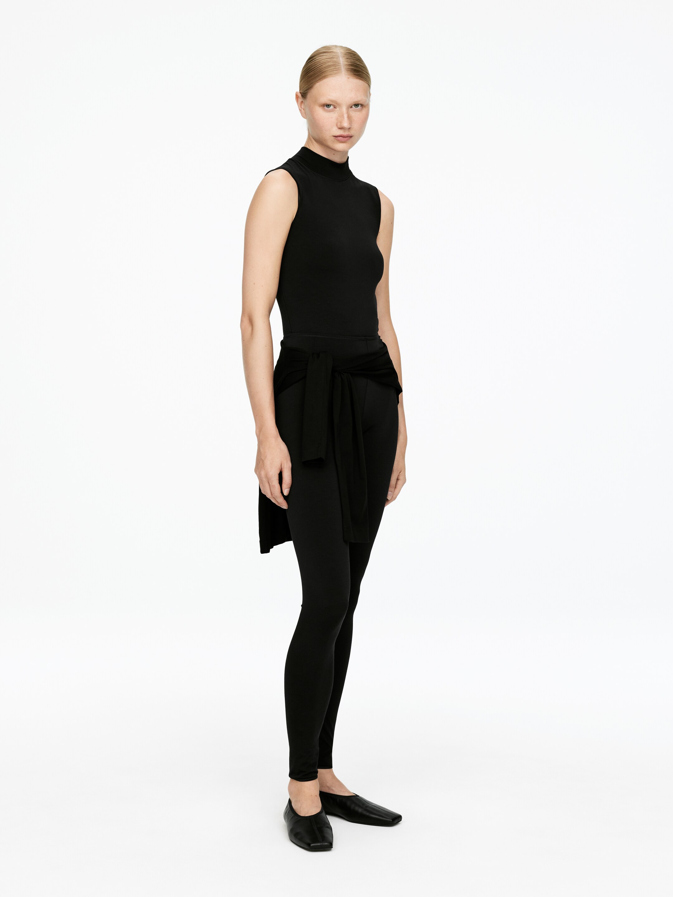 Agrandir l'image: Un haut noir sans manches à col montant est porté avec des leggings noirs moulants, créant un look élégant et superposé. Un vêtement noir assorti est noué autour de la taille, ses extrémités drapant sur les hanches et le haut des cuisses, tandis que des chaussures plates à bout carré noires complètent l'ensemble.