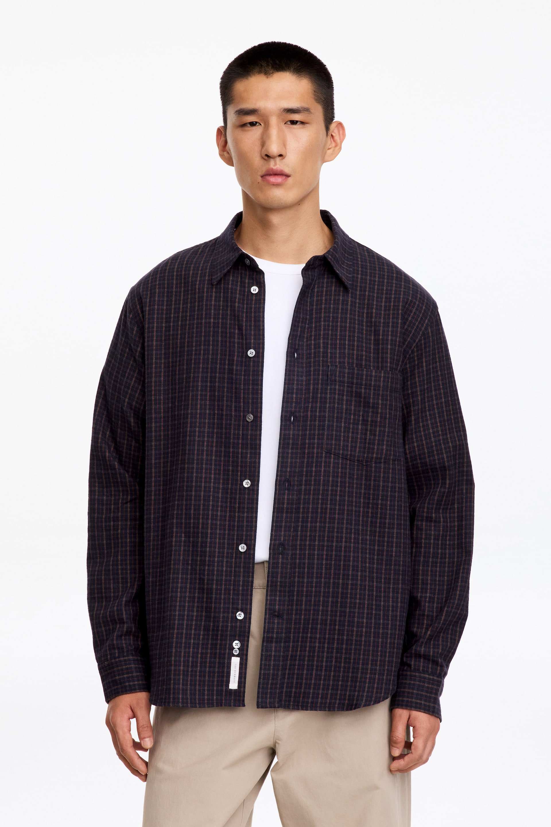 Visualizza immagine più grande: Una camicia di flanella a fantasia a quadri scuri è indossata sbottonata dal modello sopra una t-shirt bianca ed è abbinata a pantaloni beige. La camicia dalla vestibilità rilassata presenta un colletto, una singola tasca applicata sul petto e bottoni chiari.