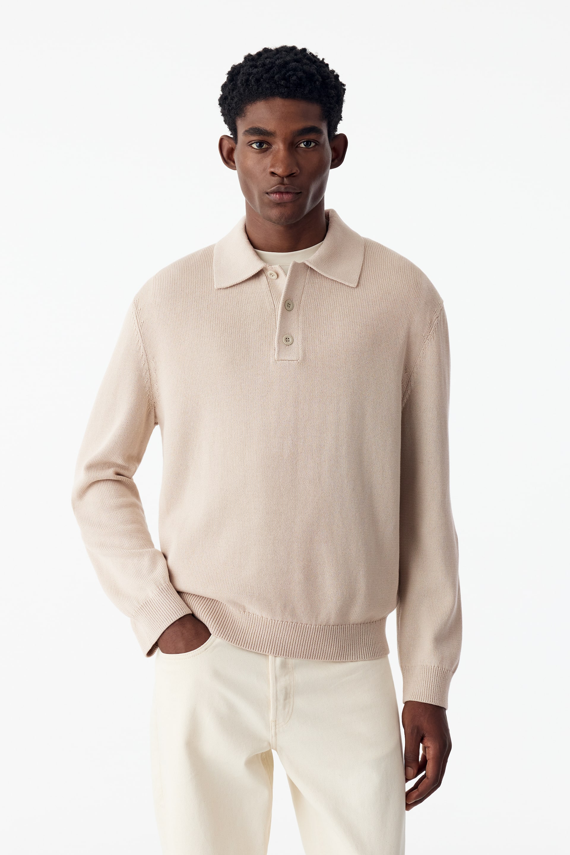 Ver imagem maior: Uma polo de malha fina em bege-claro está a ser usada pela modelo, apresentando mangas compridas, gola clássica, carcela curta com três botões e bainha e punhos canelados. A polo de corte descontraído é usada por cima de uma t-shirt branca e combinada com calças branco-sujo.