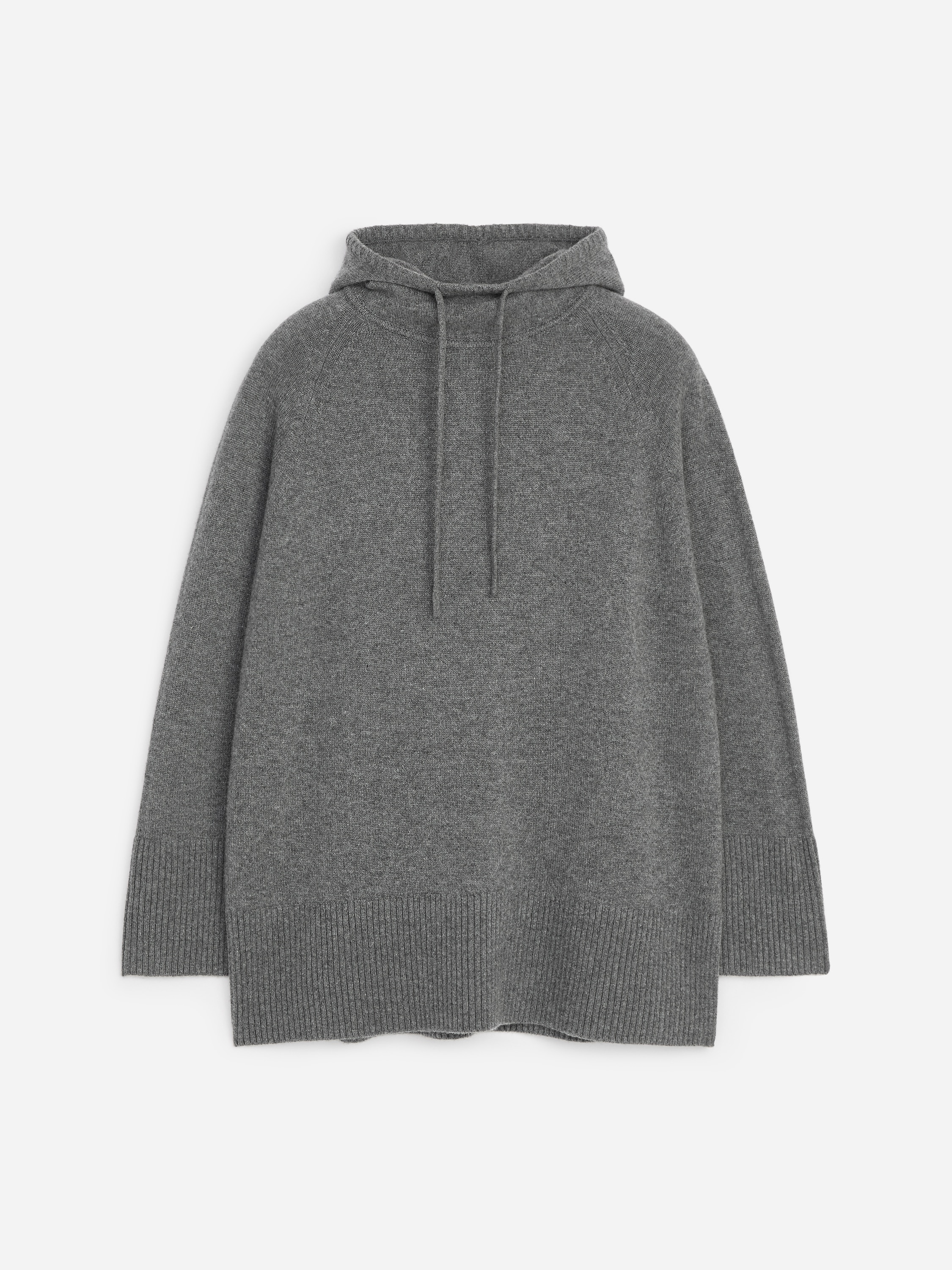 En grÃ¥ kashmirhettegenser med lang silhuett bÃ¦res av modellen, og har justerbar hette med snÃ¸ring, raglanermer og brede ribbestrikkede kanter ved mansjettene og nederkanten. Hettegenseren styles med matchende grÃ¥ strikket bukse.