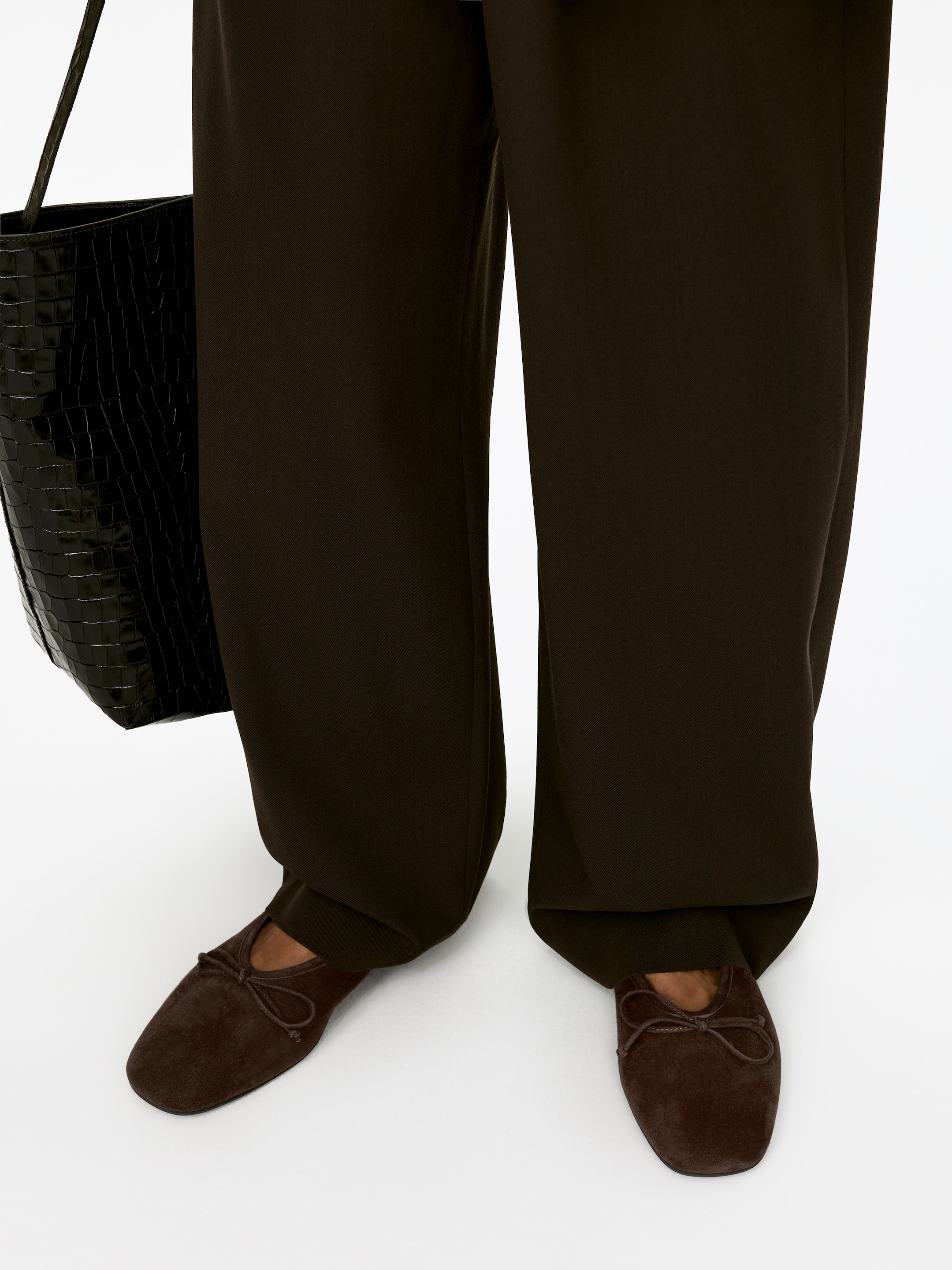 Visualizza immagine più grande: Classiche ballerine in camoscio marrone scuro sono indossate dalla modella. Le scarpe presentano una linea superiore alta e un dettaglio a fiocco sul davanti, con pantaloni a gamba larga che coprono parzialmente la loro parte superiore.