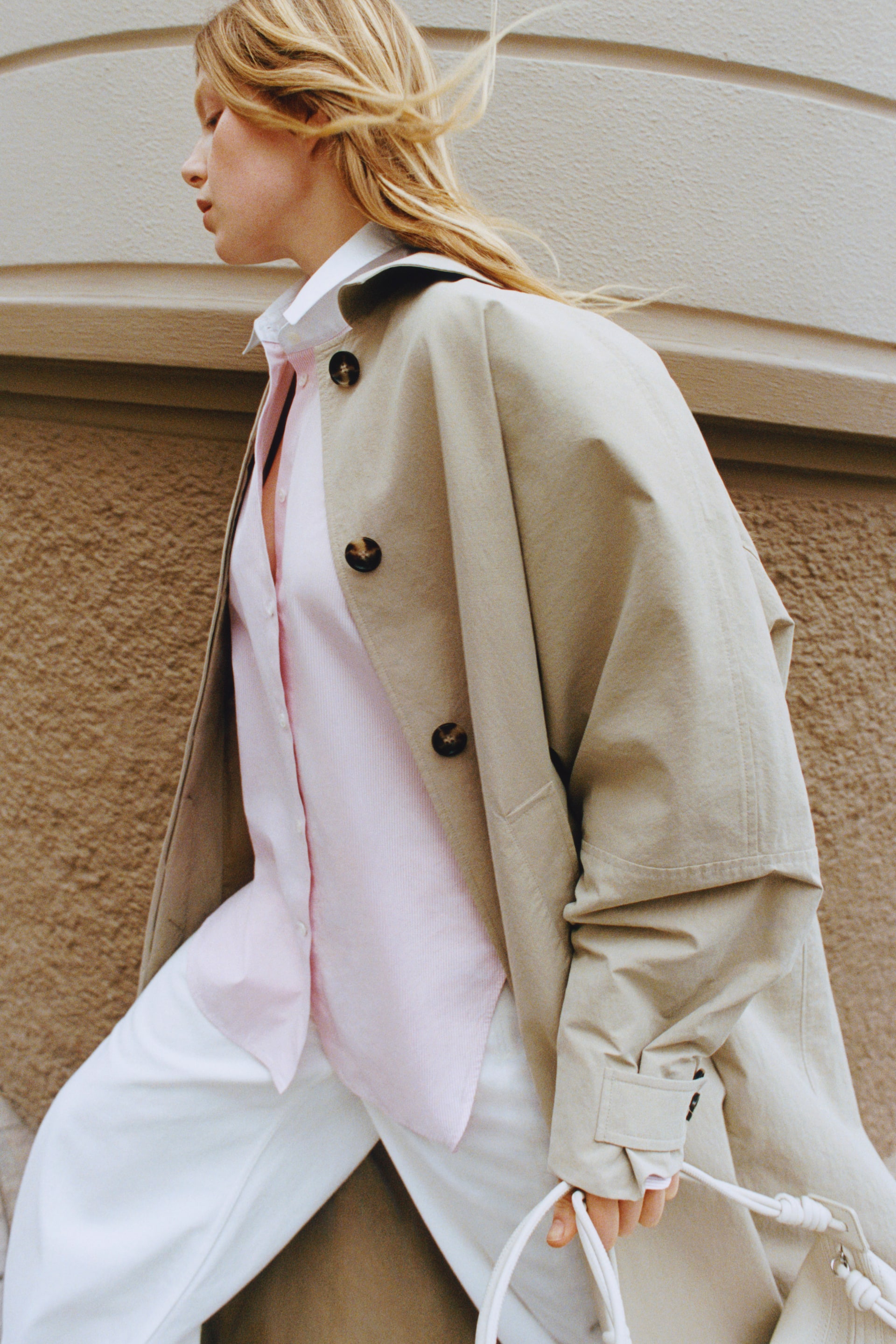 Grotere afbeelding bekijken: Een beige trenchcoat wordt gepresenteerd vanuit een zij-voorhoek, met grote donkerbruine knopen aan de voorkant en een tab met een knoop op de manchet, terwijl hij open wordt gedragen over een lichtroze gestreept shirt.