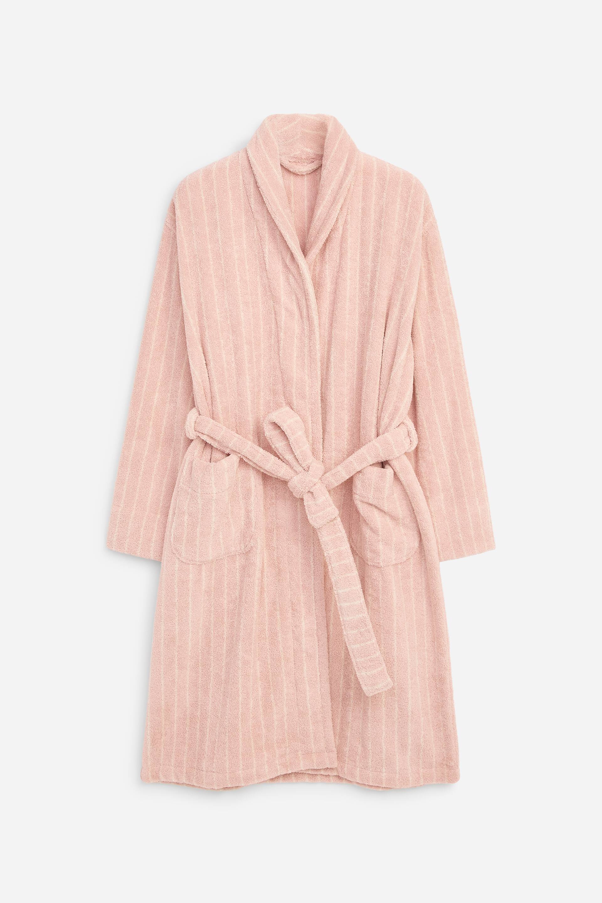 View larger image: A front-facing light pink terry towelling bathrobe is displaying a subtle vertical stripe pattern, featuring a shawl collar, long sleeves, two front patch pockets, and a matching self-tie belt knotted at the waist.