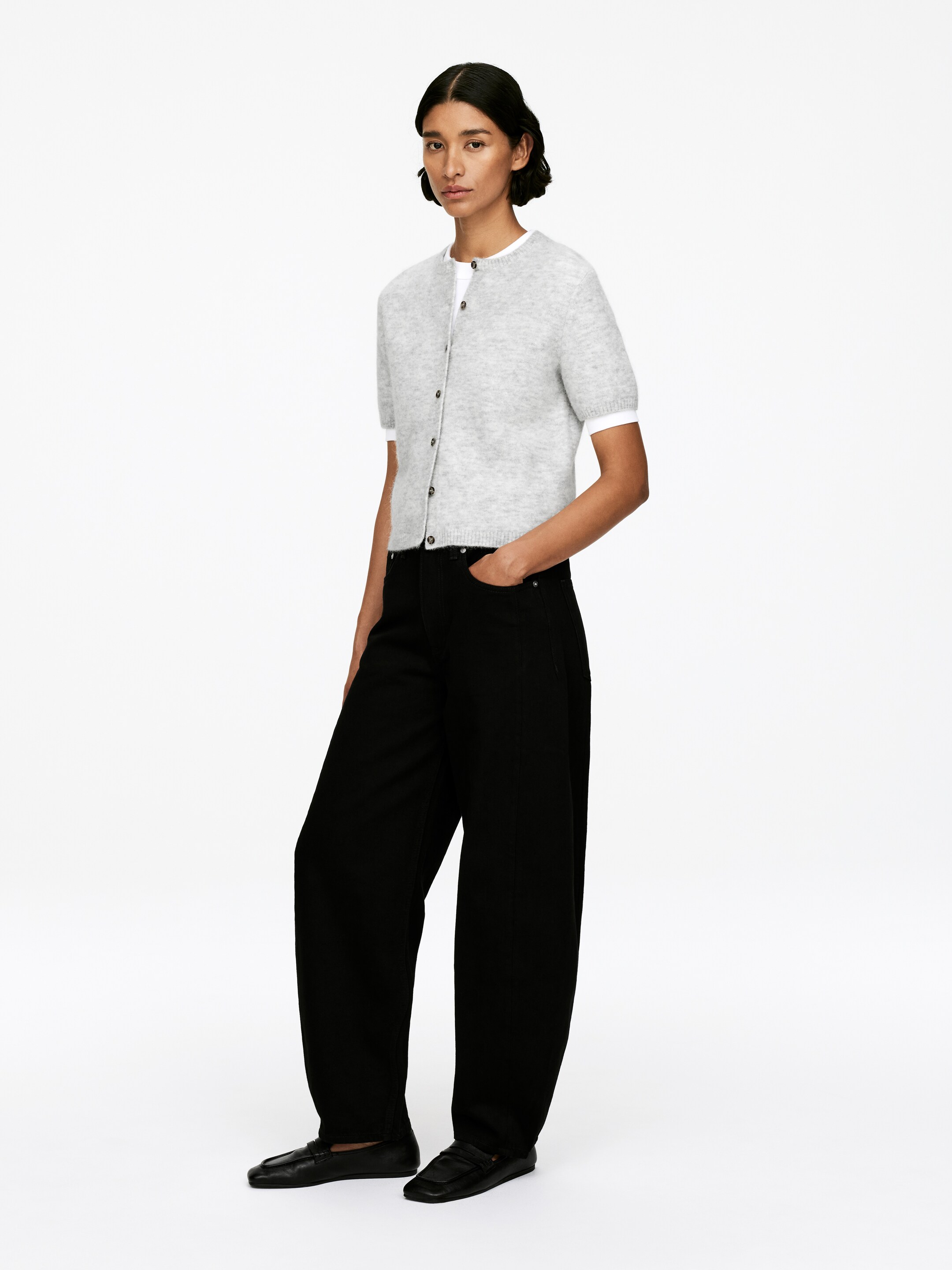 Grotere afbeelding bekijken: Een lichtgrijs kortmouwig vestje wordt gepresenteerd, dichtgeknoopt over een wit kortmouwig topje, met een ronde halslijn en geribde zoom. Het vestje wordt gecombineerd met zwarte wijde broekspijpen en zwarte loafers met vierkante neus, allemaal getoond vanuit een lichte zijhoek om hun pasvorm en ontwerp te benadrukken.