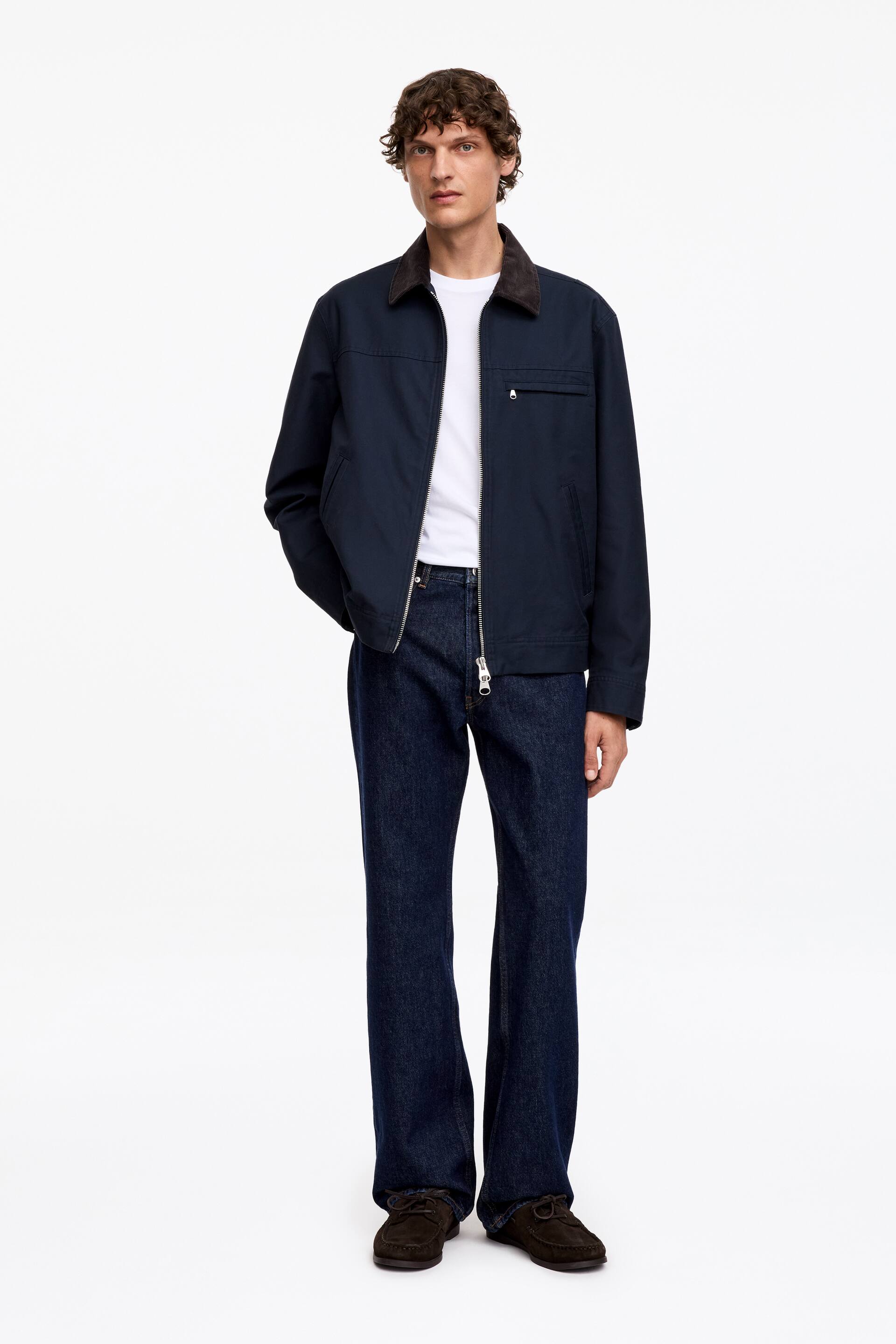Agrandir l'image: Une veste en toile de coton bleu marine, avec un col en velours côtelé marron, deux poches passepoilées à l'avant, une poche poitrine zippée et des poignets avec fermetures à boutons-pression, est portée par le mannequin. Sa fermeture éclair métallique à double sens est partiellement ouverte, révélant un t-shirt blanc en dessous, et le mannequin glisse ses deux mains dans les poches passepoilées de la veste.
