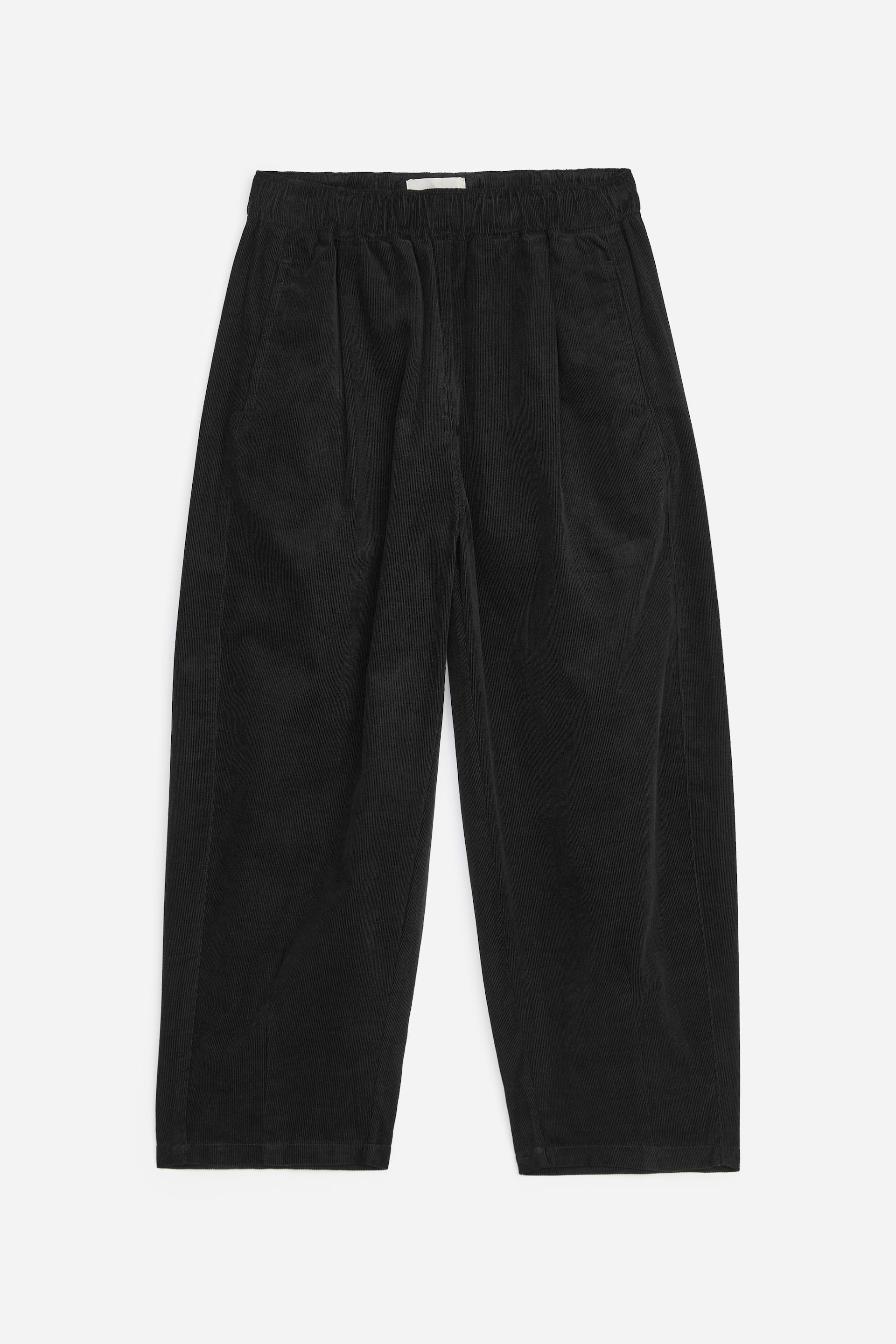 Vezi imaginea mai mare: Pantalonii negri din catifea reiată sunt prezentați din față, având talie elastică, două pense pe fiecare parte în față și buzunare în cusătura laterală. Pantalonii au, de asemenea, croială largă a piciorului și tiv drept.