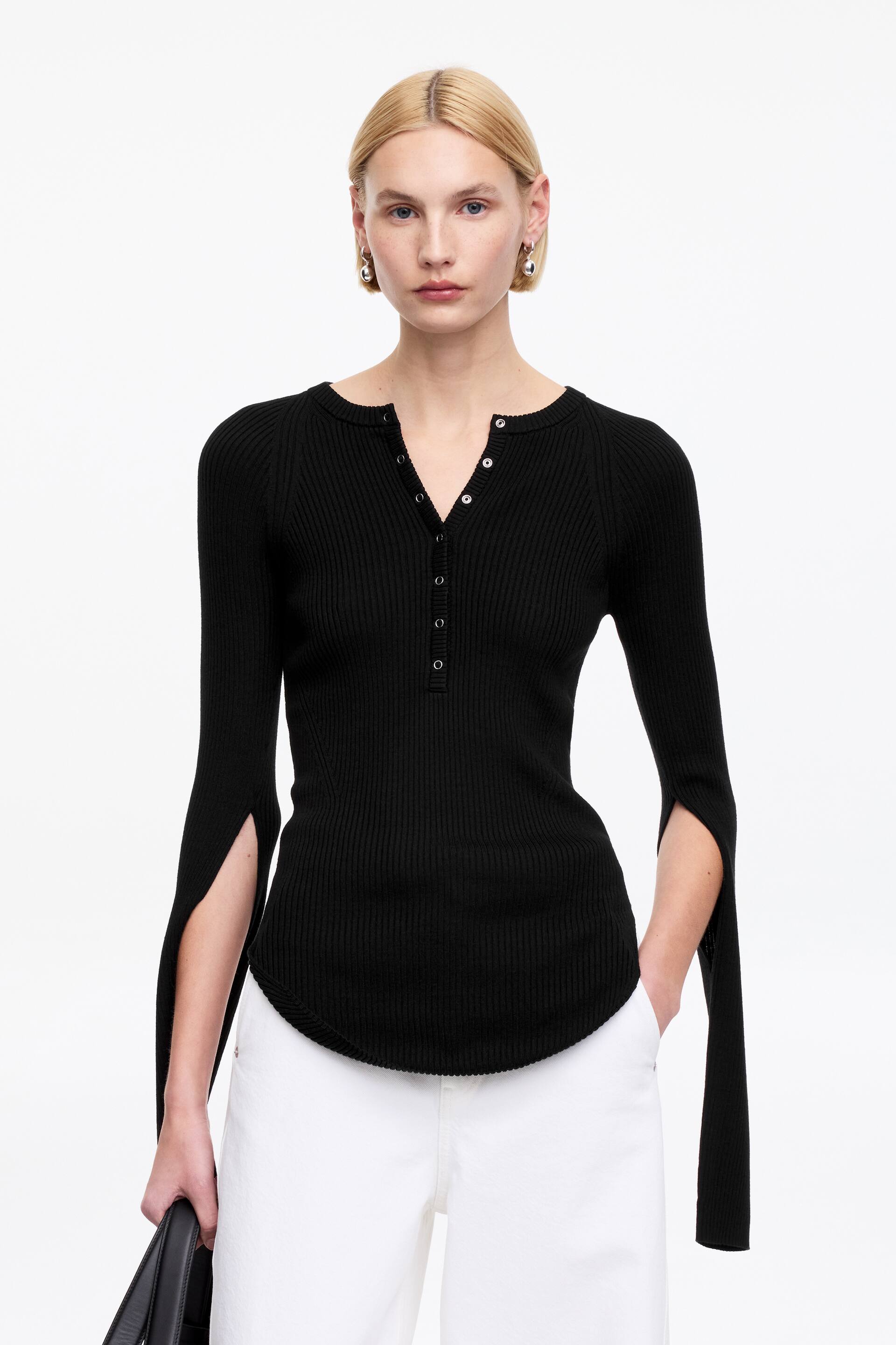 Vis større bilde: En svart ribbestrikket topp med en Henley-utringning og en fem-knappers stolpe. Den har lange ermer med splittede mansjetter som eksponerer underarmene, samt en avrundet kant som skaper en smal silhuett.