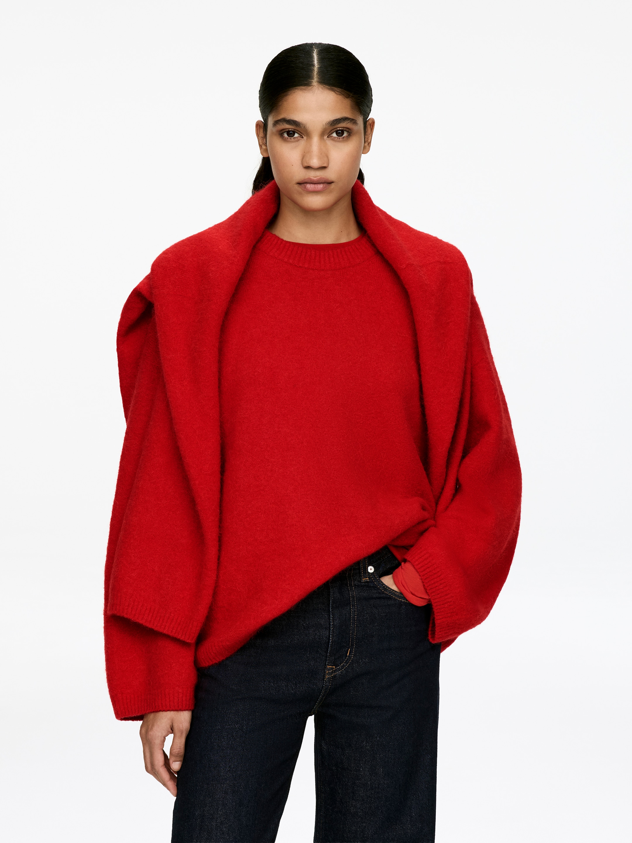 Agrandir l'image: Un pull à col rond rouge est porté, présentant une coupe décontractée, légèrement surdimensionnée, des poignets côtelés visibles et une texture douce et duveteuse. Un deuxième vêtement rouge est drapé sur les épaules, créant un effet superposé.