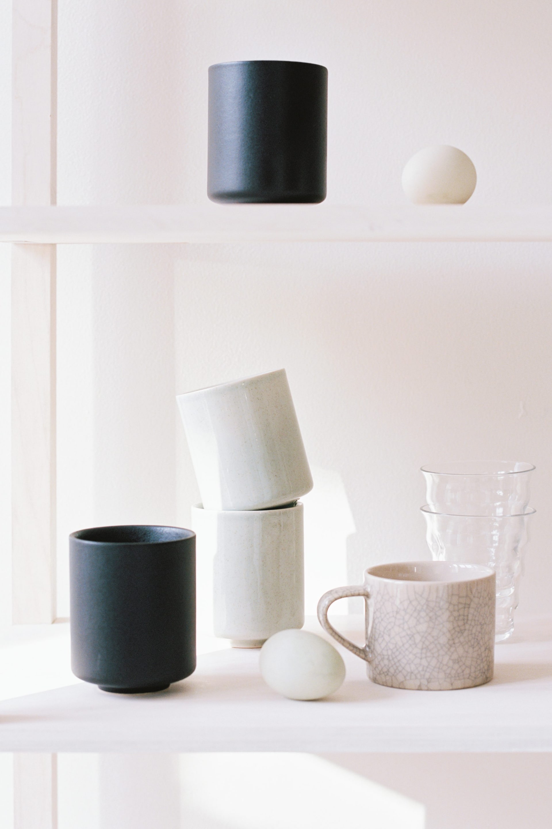 Vis større bilde: En samling av keramiske og glasskar er utstilt, med en matt svart sylinderformet kopp, en annen matt svart sylinderformet kopp med en bredere base, to lysegrønne sylinderformede kopper stablet i en vinkel, og et lyst keramikkrus med krakelert glasur og håndtak. To klare glasskrus med en uregelmessig teksturert overflate er også stablet.