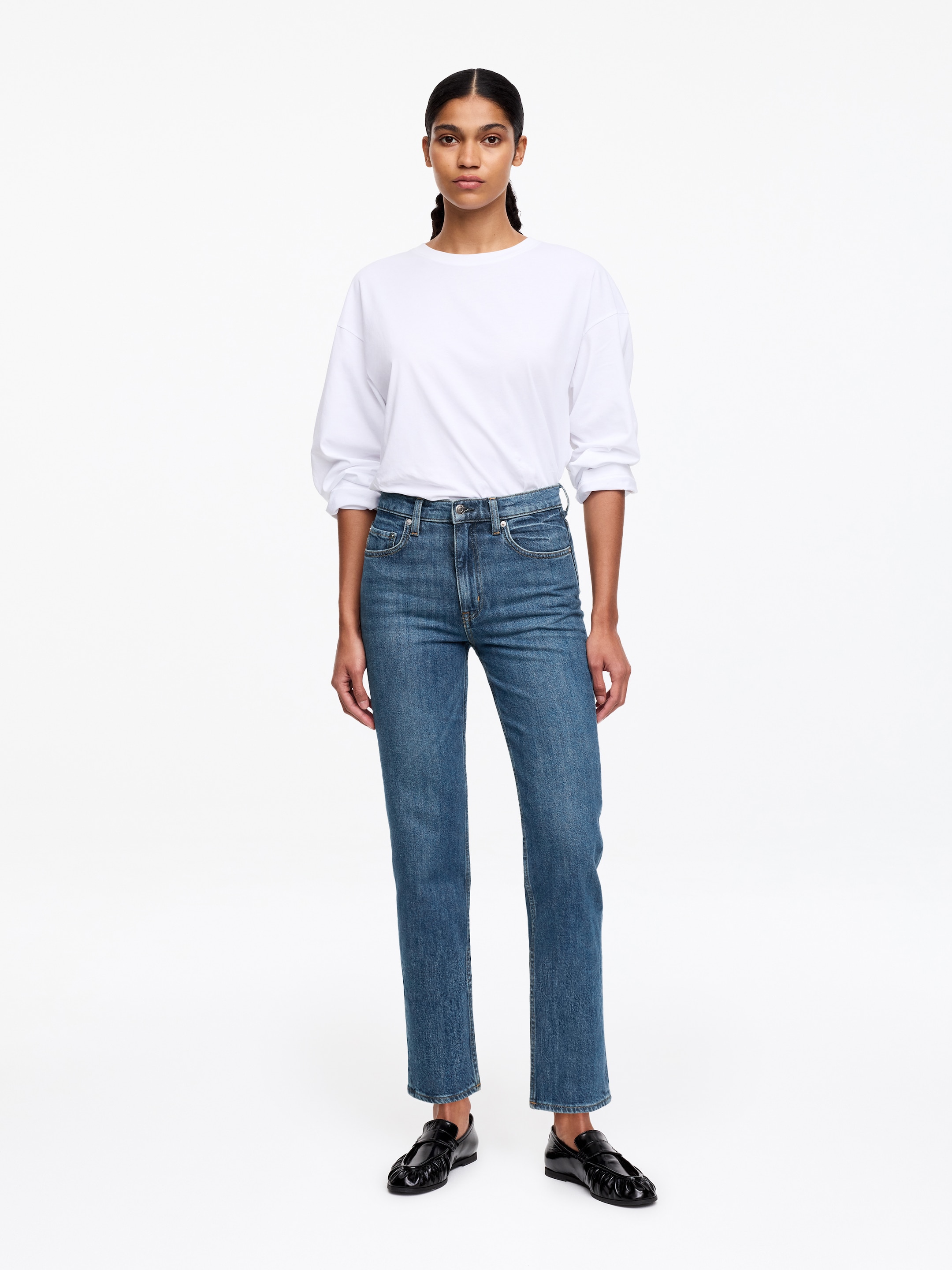 Grotere afbeelding bekijken: Een paar middelblauwe, hoog getailleerde, recht gesneden jeans met een subtiele vervaagde wassing en een cropped lengte, met klassieke vijfzakken styling. De jeans worden gepresenteerd met een wit, lang mouw crew-neck t-shirt, dat nonchalant aan de voorkant is ingestopt.