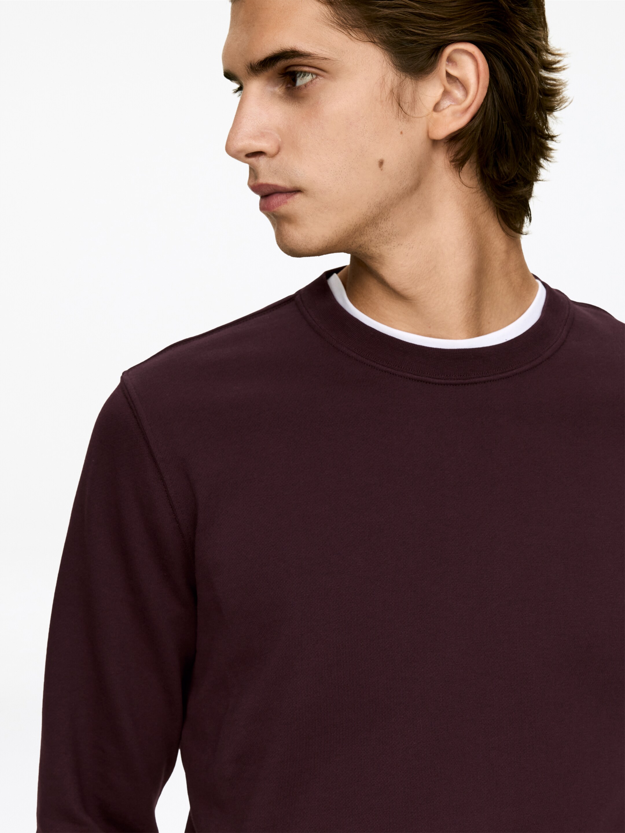Ver imagen más grande: Se muestra una sudadera color granate con cuello redondo desde una vista lateral parcial, mostrando mangas largas y una textura de tela lisa. Se ve un cuello de camiseta blanca debajo de su cuello redondo.