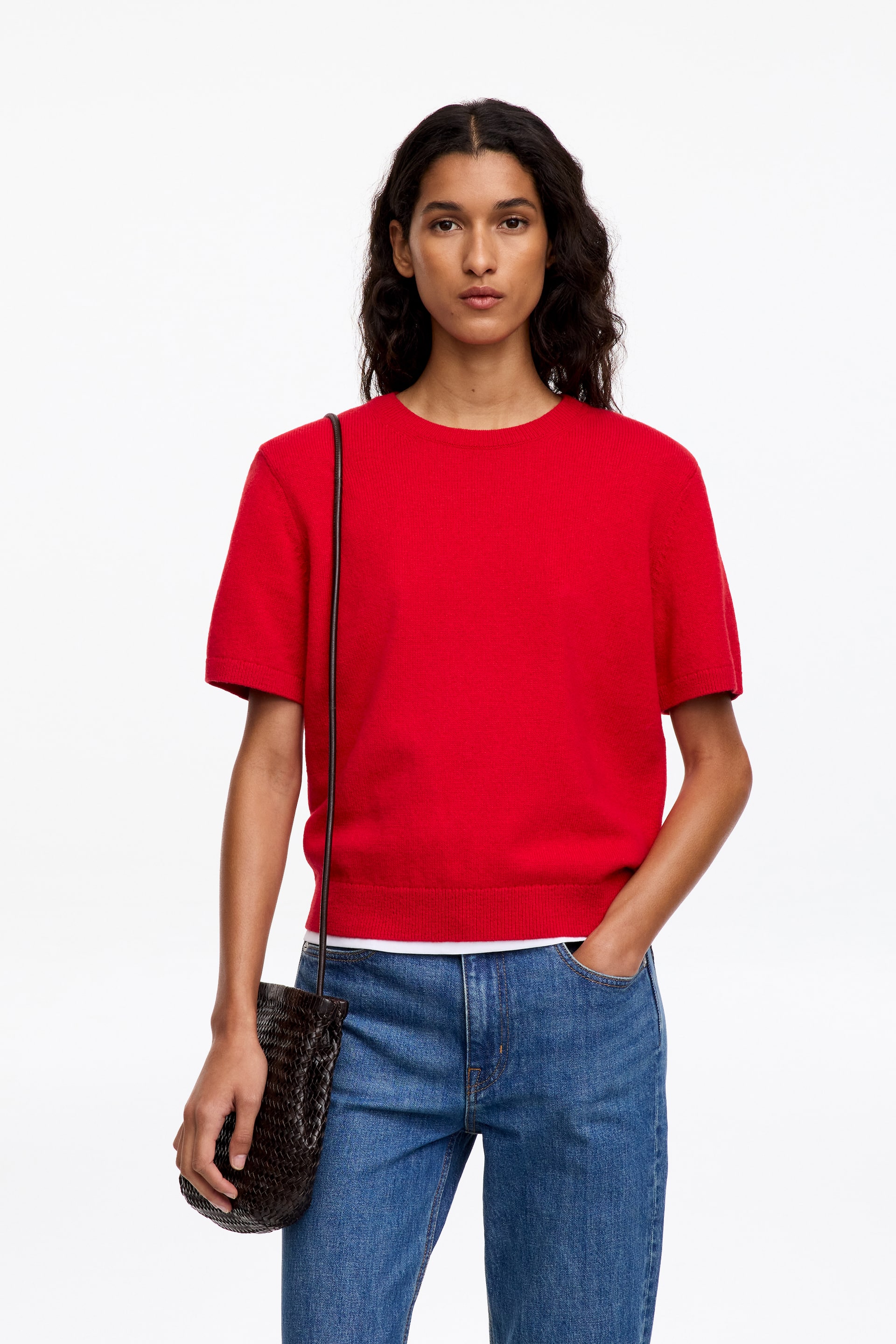 Grotere afbeelding bekijken: Een rode, gebreide trui met korte mouwen, ronde hals en geribde boorden wordt gedragen door het model. De trui wordt gedragen over een wit T-shirt en gestyled met een blauwe jeans en een donkerbruine geweven schoudertas.