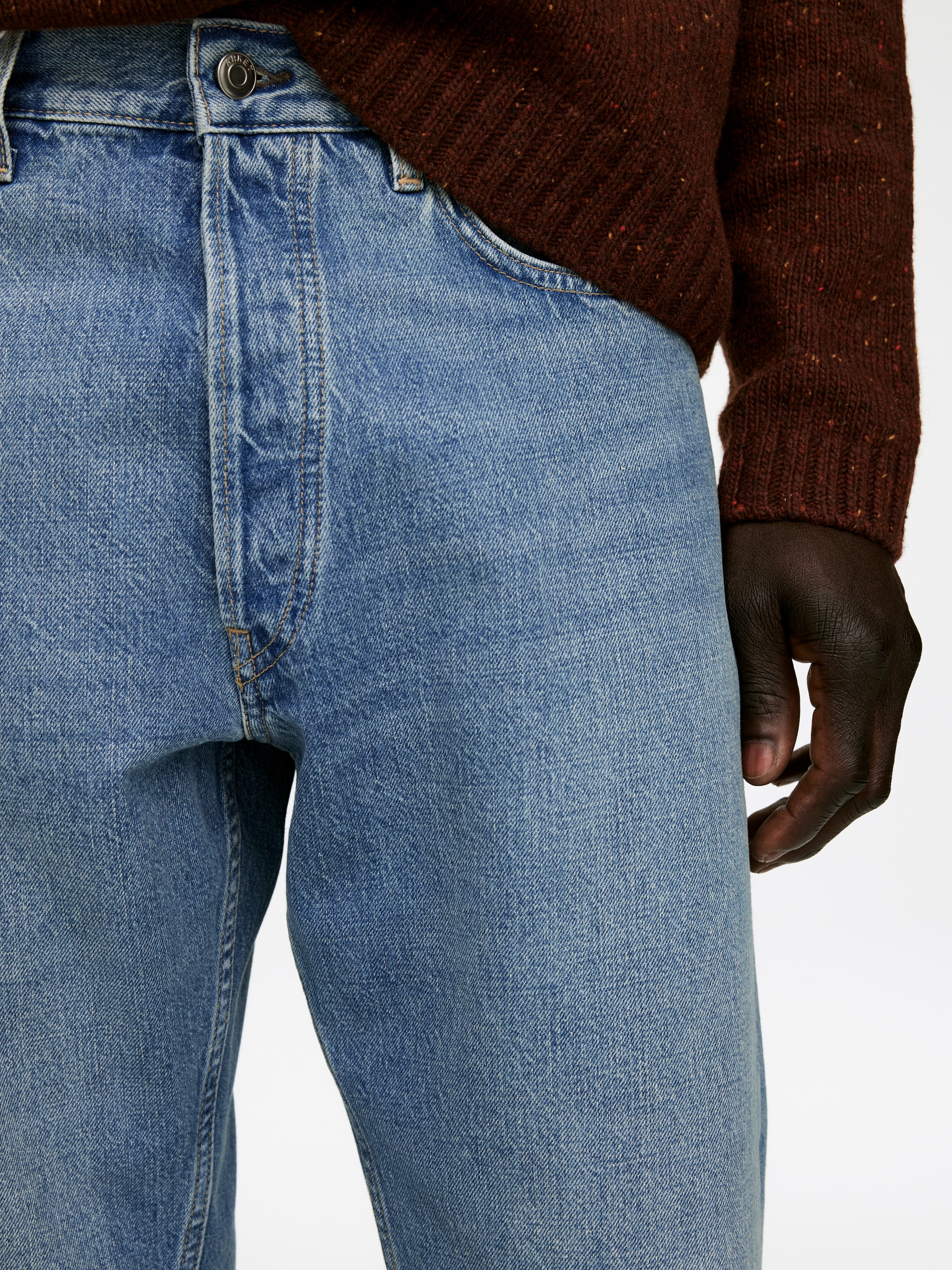 Grotere afbeelding bekijken: Licht gewassen blauwe denim jeans hebben een zichtbare knoopsluiting, riemlussen en ronde voorzakken, zoals te zien is vanaf de voorkant onder een lichte hoek. De jeans met een ontspannen pasvorm worden gedragen met een bruine gebreide trui.