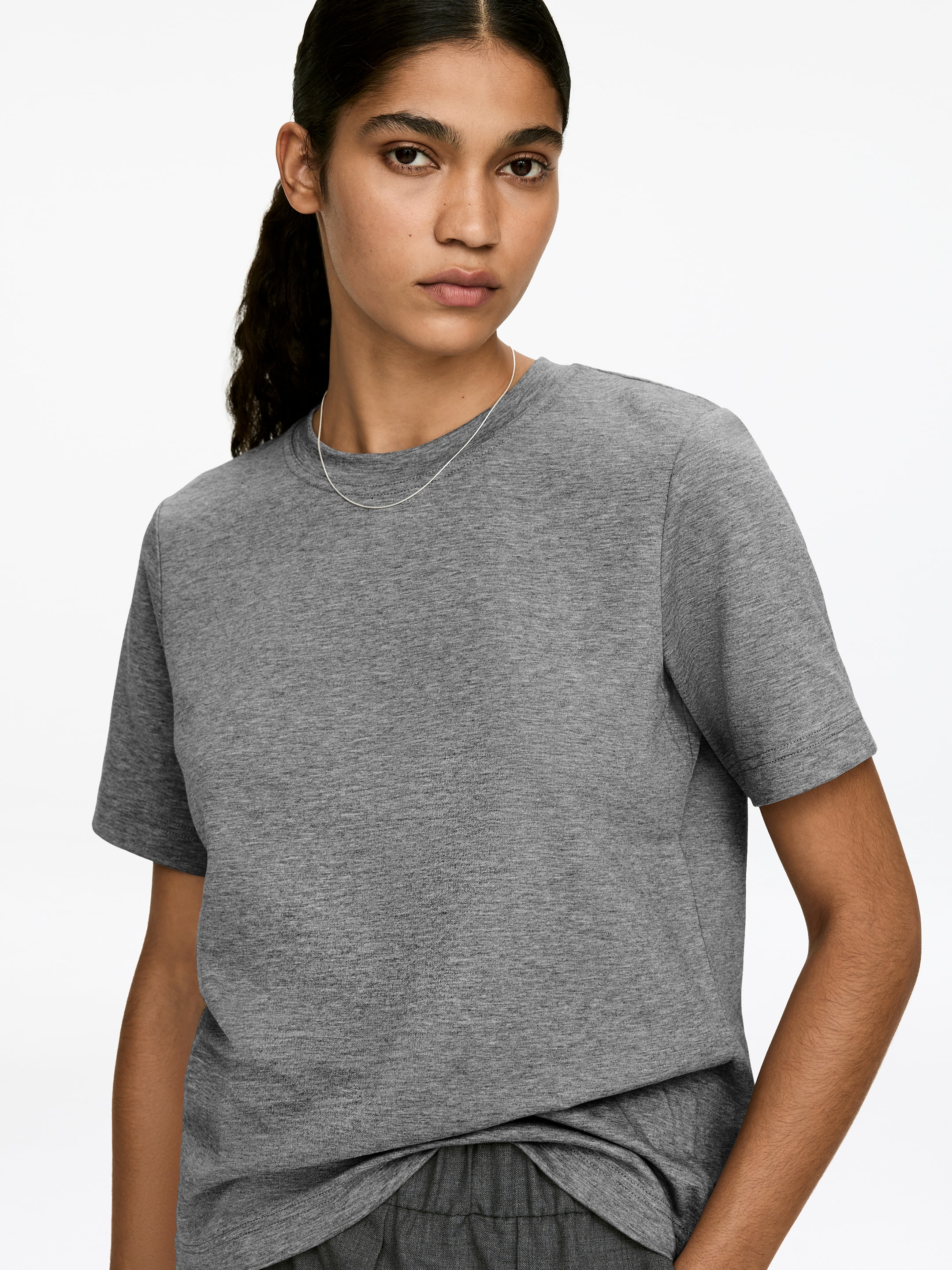 View larger image: A grey T-shirt with a classic crew neck and short sleeves is being worn by the model. The regular-fit T-shirt, featuring a heathered jersey texture, is styled with a thin silver necklace resting over it and dark grey pants visible beneath its hem.