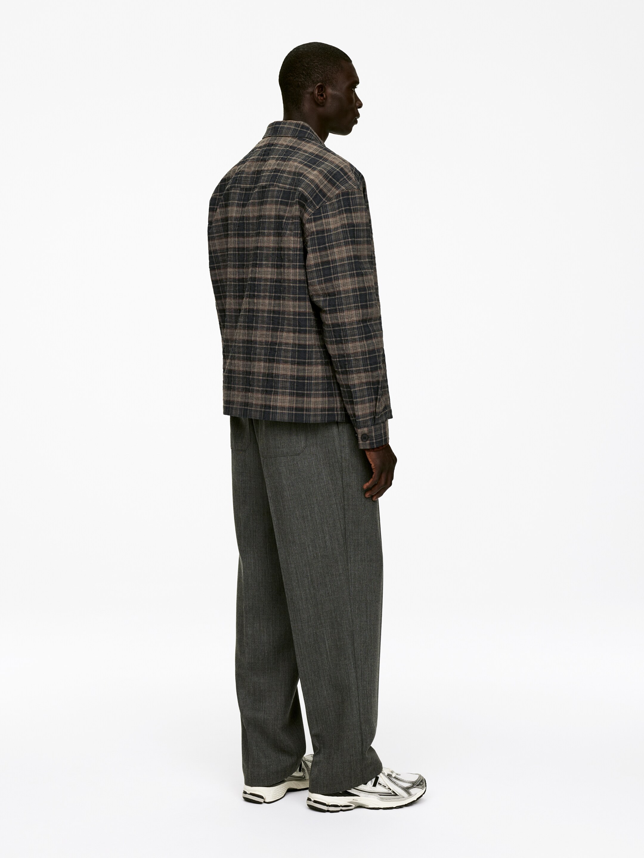 Agrandir l'image: Une chemise-veste à carreaux de couleur charbon foncé et beige est portée, sa coupe ample visible de profil. Elle est superposée à un pantalon gris foncé à jambes larges avec des rayures verticales subtiles. L'ensemble complet est complété par des baskets athlétiques blanches, argentées et noires.