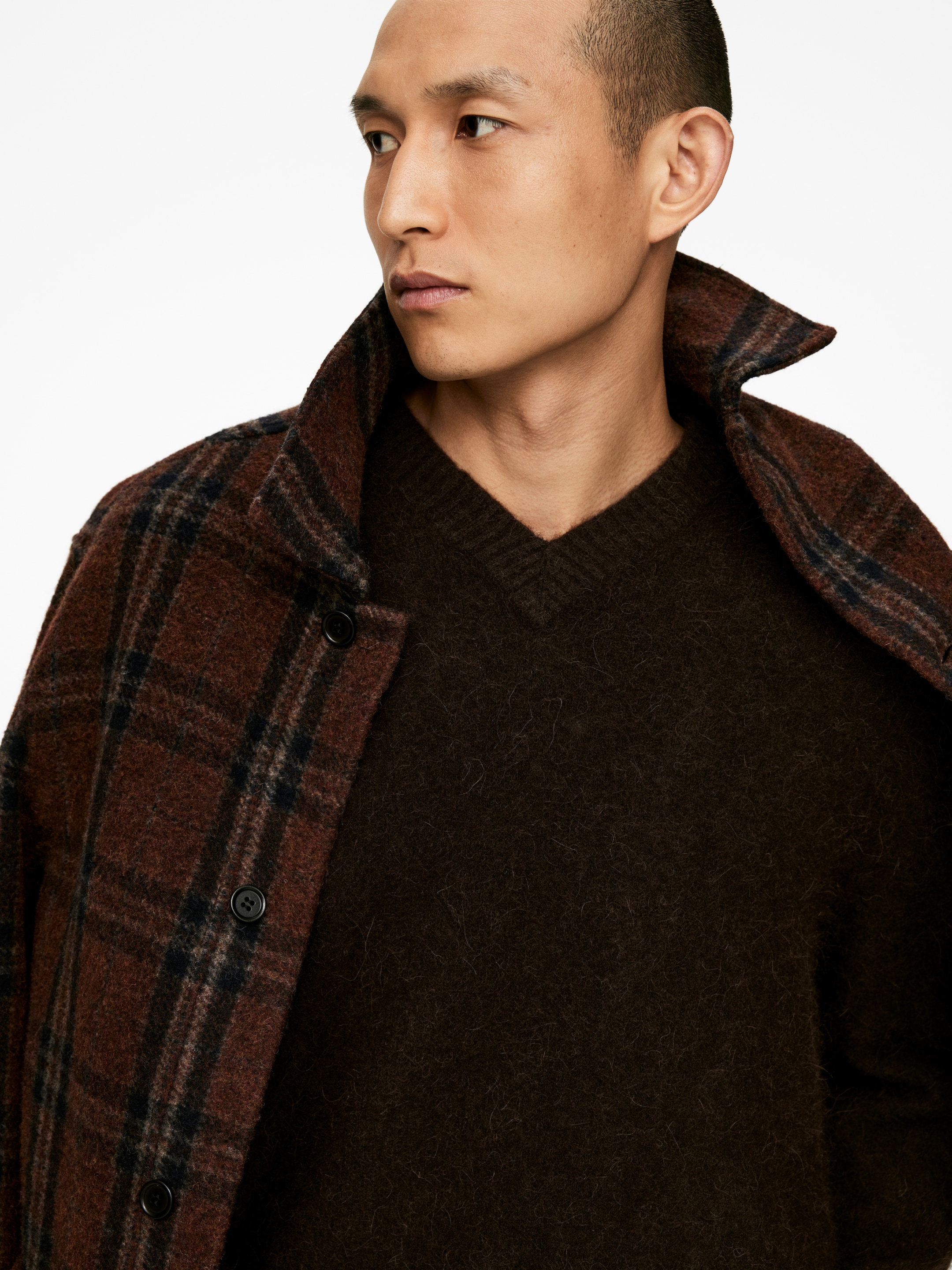 Agrandir l'image: Une veste à carreaux marron est portée ouverte sur un pull à col en V marron foncé. La veste présente un col montant et des boutons noirs sur sa patte de boutonnage avant, tandis que le pull arbore une texture douce et duveteuse.