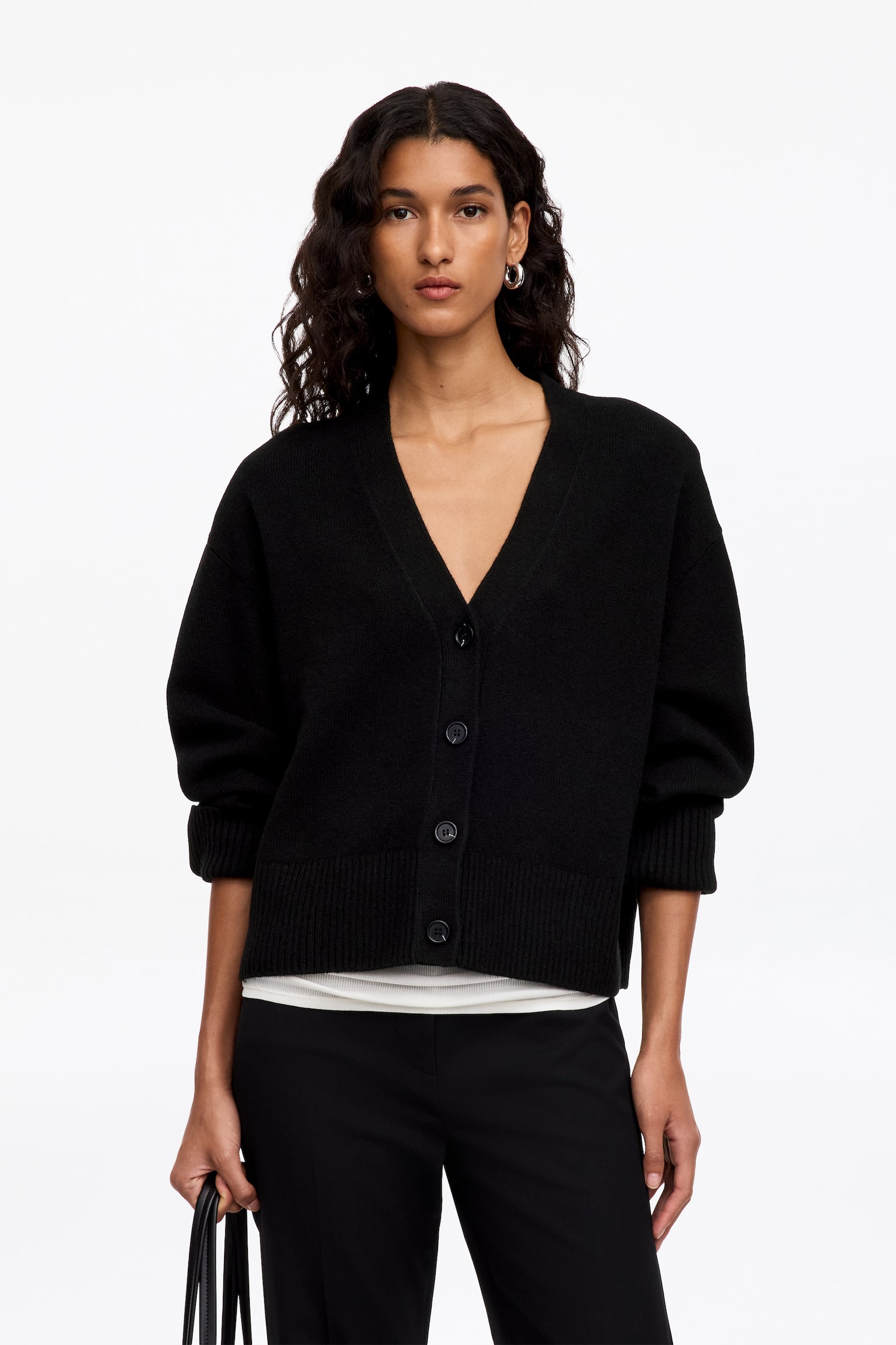 Una donna indossa un cardigan nero in cashmere con scollo a V profondo, chiusura frontale con bottoni e bordi a coste su polsini e orlo. Il cardigan è abbinato a un top bianco e a pantaloni neri. - 1