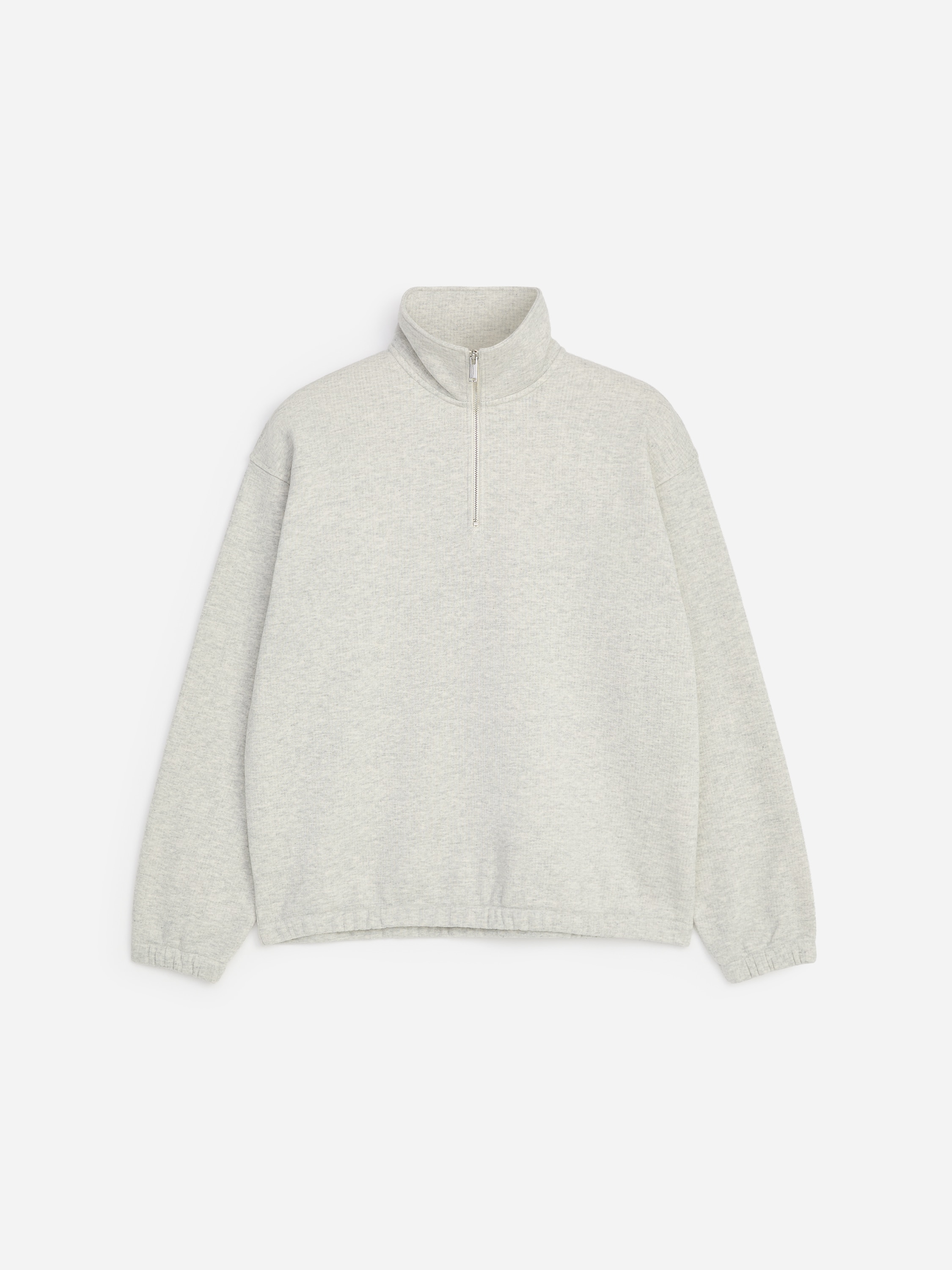 A modelo está a usar uma sweatshirt cinzento-claro com meio fecho, gola alta, ombros descaÃdos e punhos e bainha elásticos. A sweatshirt está sobreposta a uma t-shirt branca e combinada com uns calções de lazer cinzento-claro a condizer.