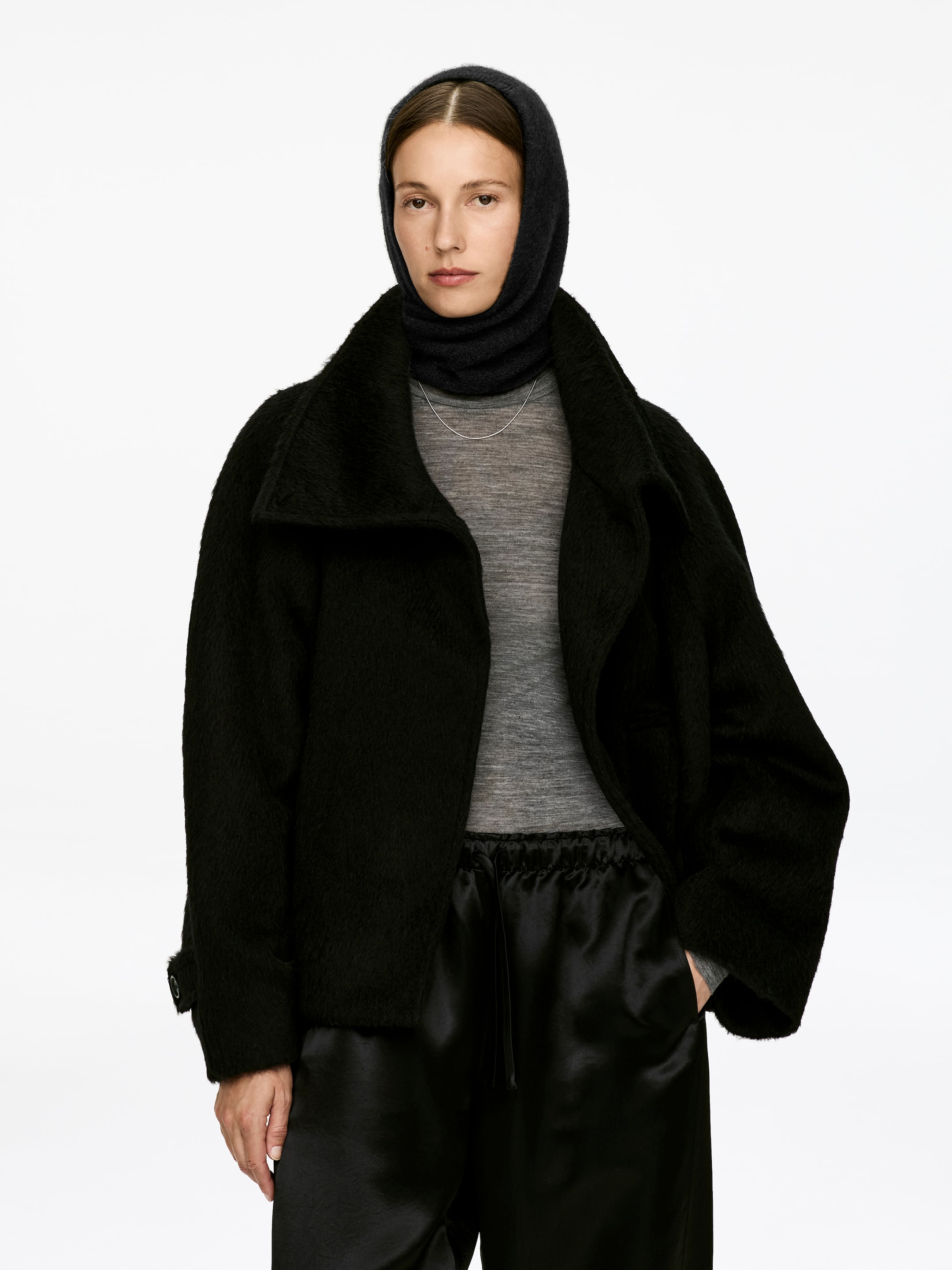 Ver imagen más grande: Una capucha oscura y texturizada, elaborada con una mezcla de alpaca y lana, está siendo llevada por la modelo. Cubre su cabeza y cuello, enmarcando su rostro, y está superpuesta sobre un top gris y debajo de una chaqueta negra.