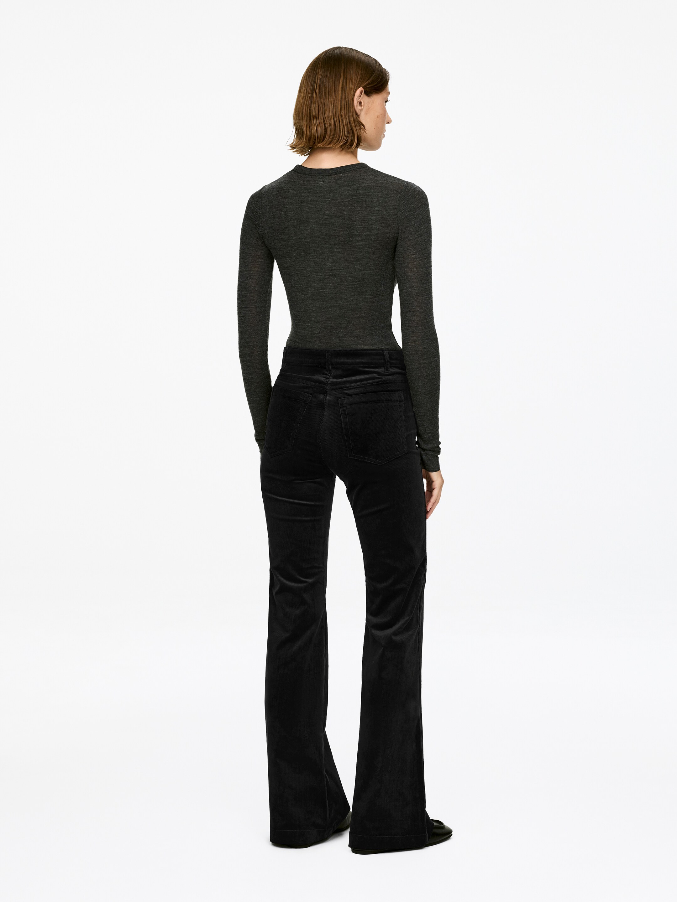 Ingrandisci l'immagine: Pantaloni neri in velluto a coste, caratterizzati da una vita alta, una vestibilità aderente attraverso la coscia e gambe svasate, sono indossati dalla modella. Una maglia scura a maniche lunghe è indossata infilata nei pantaloni, e scarpe basse scure sono indossate sotto gli orli svasati.