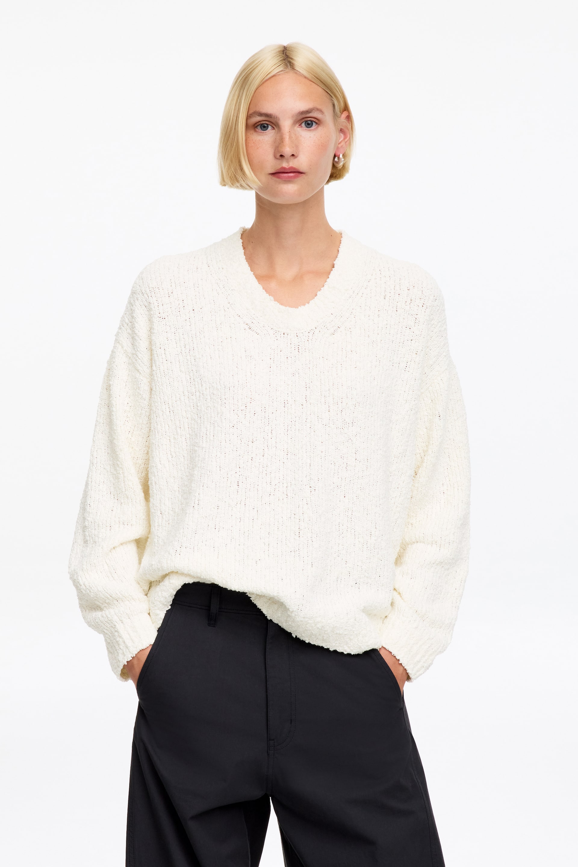 Agrandir l'image: Un pull crème à col en V est présenté, avec un tricot texturé et des manches longues. Il est conçu dans une coupe décontractée, avec son ourlet partiellement rentré dans un pantalon foncé.
