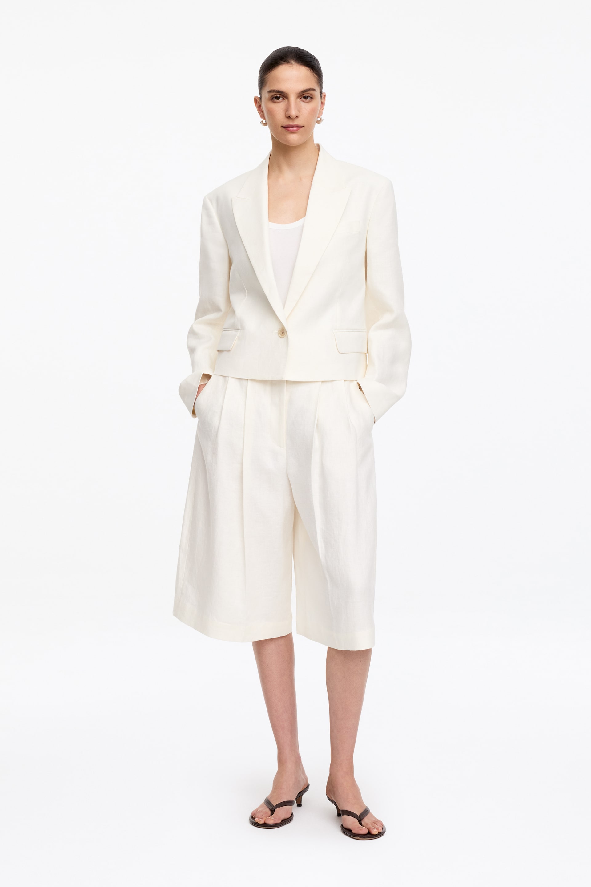Agrandir l'image: Un blazer court en mélange de lin blanc cassé et des culottes plissées assorties sont exposés. Le blazer à simple boutonnage présente des revers crantés, une fermeture à un bouton et des poches à rabat à l'avant, et est superposé sur un débardeur blanc, tandis que les culottes sont dotées de plis et de poches latérales.