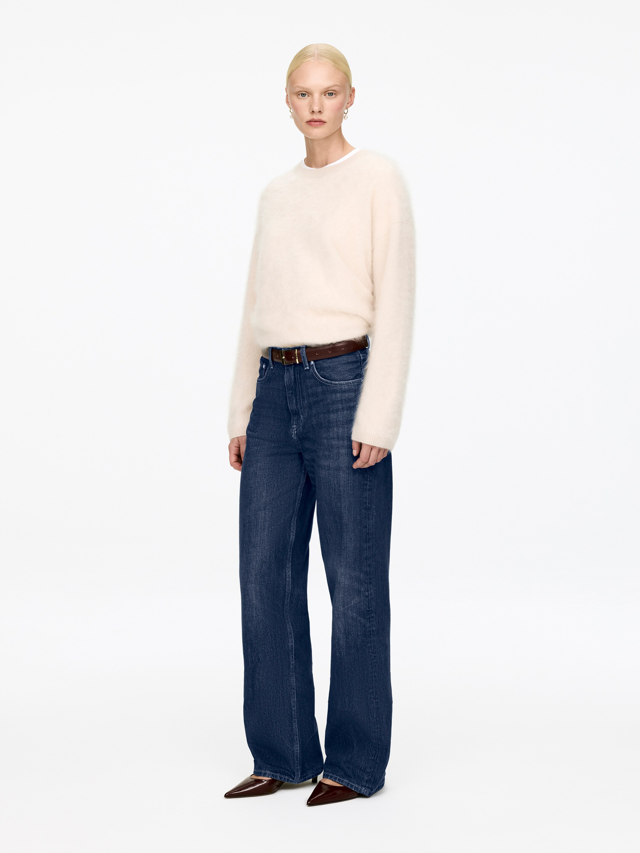 Größeres Bild anzeigen: Ein cremefarbenes, flauschiges Rundhals-Pullover wird mit dunkelblauen, weiten Jeans getragen, die nach vorne gerichtet präsentiert werden. Die Jeans werden mit einem braunen Gürtel geschnürt, und es werden dunkelbraune, spitz zulaufende Schuhe getragen.
