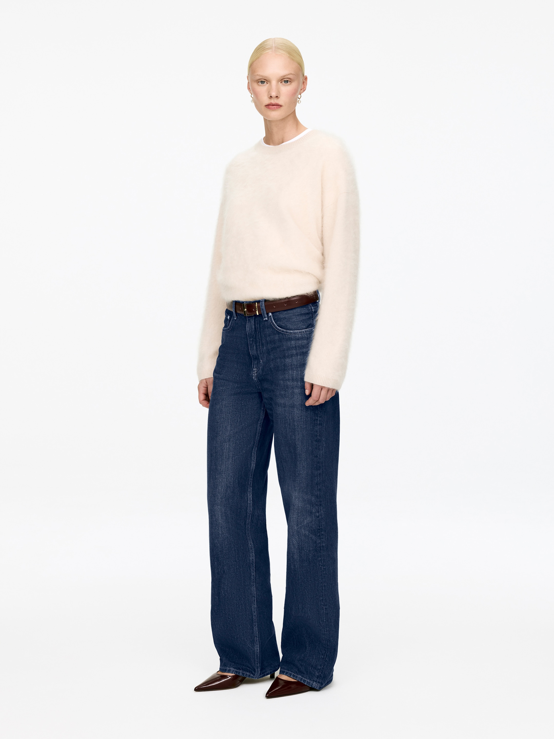 Se større billede: En cremefarvet, fuzzy crew-neck sweater bæres sammen med mørkeblå, bredbenede jeans, præsenteret med fronten mod kameraet. Jeansene bliver holdt på plads af et brunt bælte, og mørkebrune sko med spidse tå bæres.