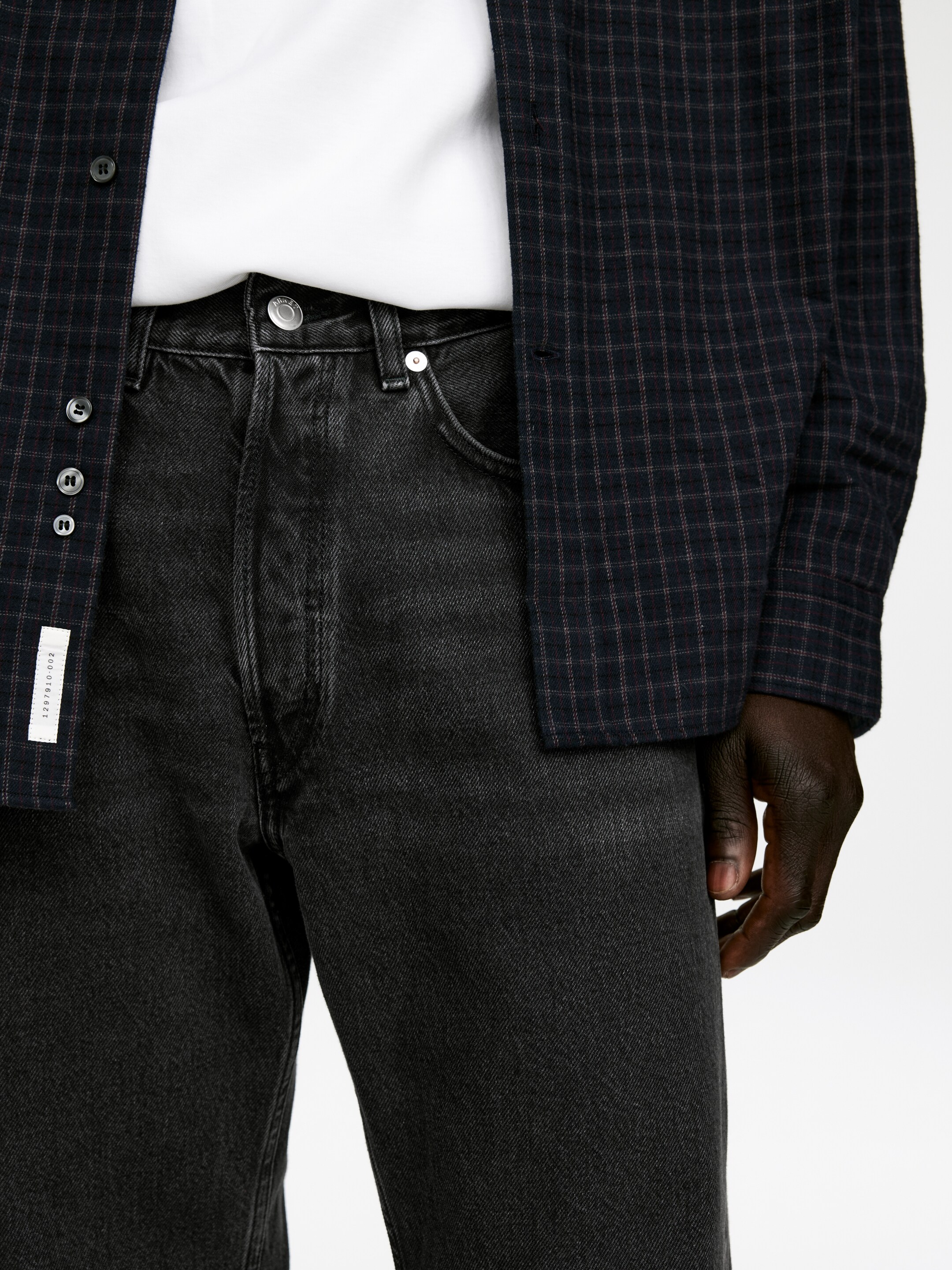 Ingrandisci l'immagine: Jeans in denim grigio scuro lavato sono indossati, con una chiusura a bottoni visibile e tasche frontali con rivetti in metallo. Una t-shirt bianca è indossata infilata nei jeans, e una camicia a quadri scura è indossata sbottonata sopra la t-shirt, parzialmente coprendo la parte superiore dei jeans.