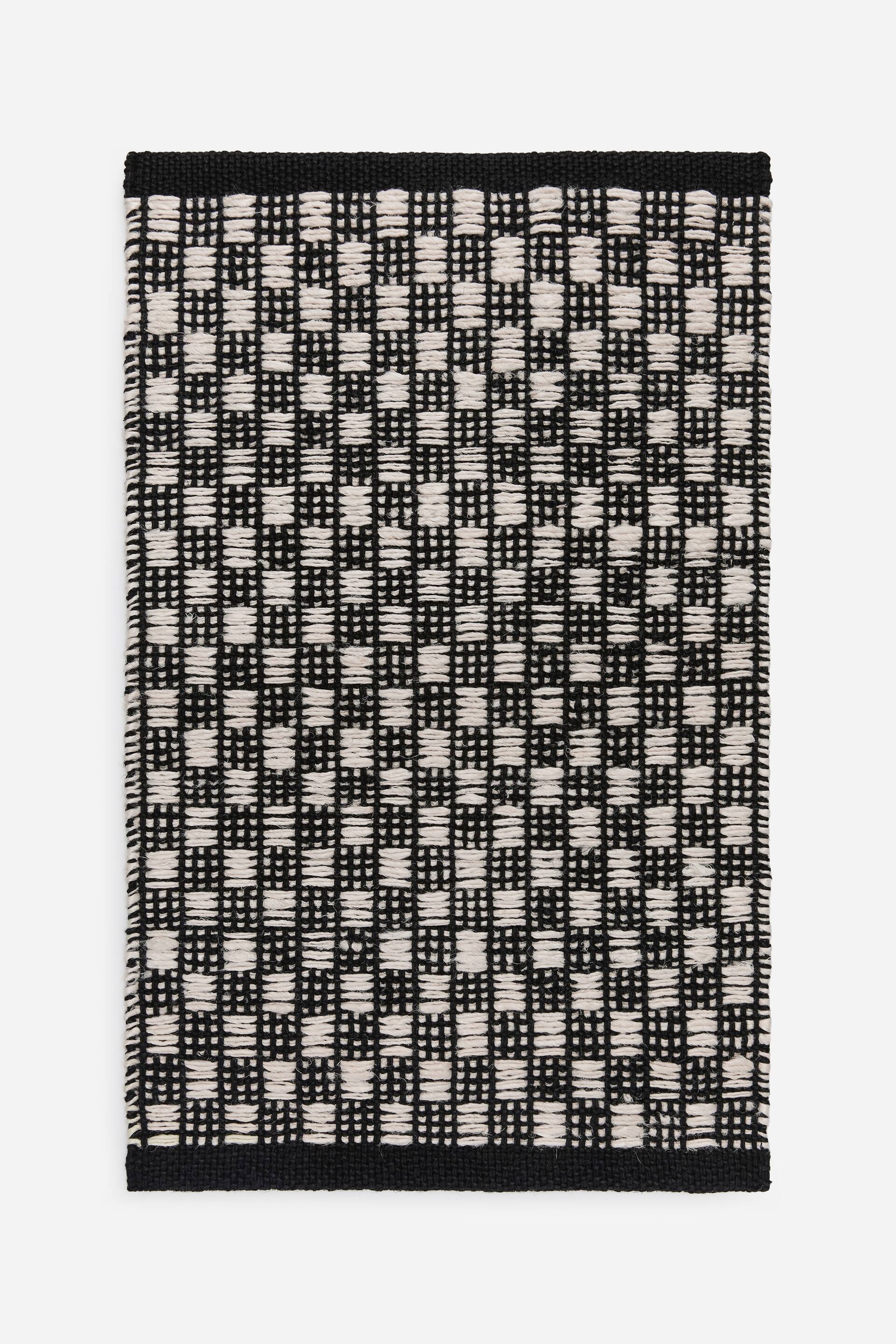 Agrandir l'image: Un tapis rectangulaire tissé présente un motif en damier composé de carrés noirs et blanc cassé alternés, avec une bande noire unie bordant ses bords supérieur et inférieur.