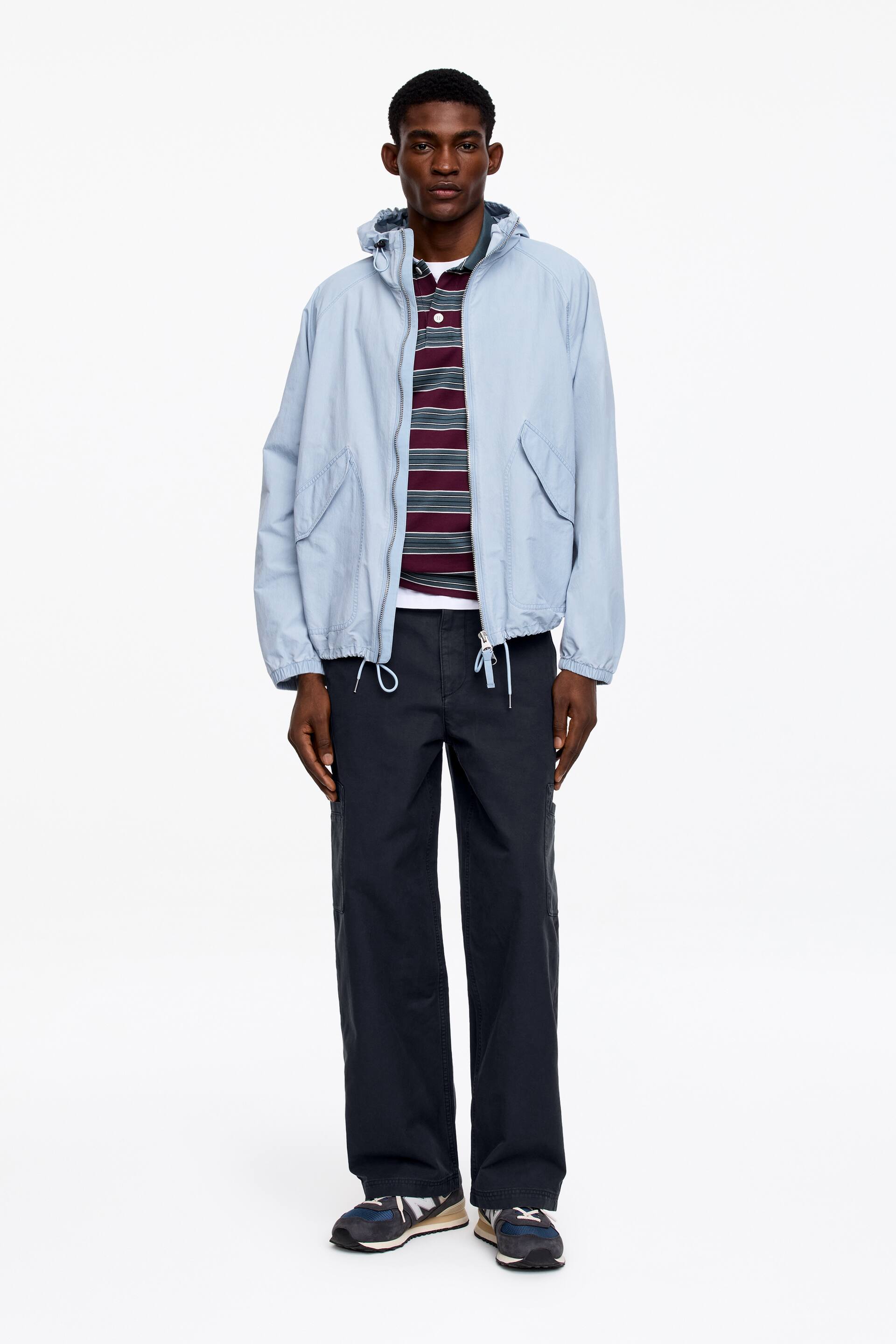 Ver imagem maior: Está a ser apresentada uma jaqueta com capuz azul-claro, com fecho de correr frontal, bolsos de abertura inclinada e bainha com cordão ajustável. Está a ser usada aberta sobre uma camisa polo às riscas em tons de bordeaux e escuro, com carcela de três botões, combinada com calças cargo largas de cor escura e ténis azul-marinho.