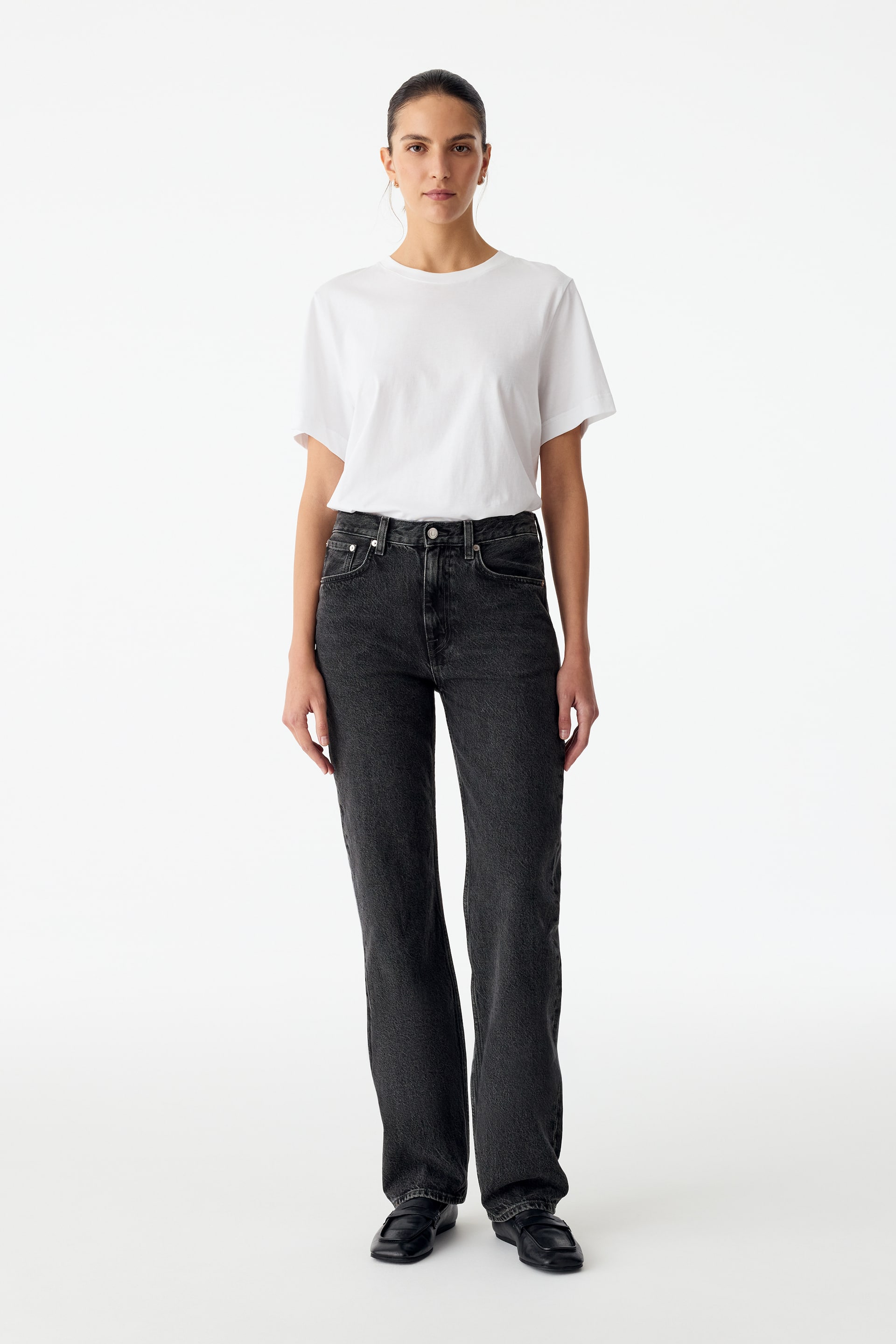 Agrandir l'image: Un jean en denim gris foncé à jambe droite et taille mi-haute est porté par le mannequin. Le jean est associé à un T-shirt blanc à manches courtes rentré dans le pantalon et à des chaussures noires à enfiler.