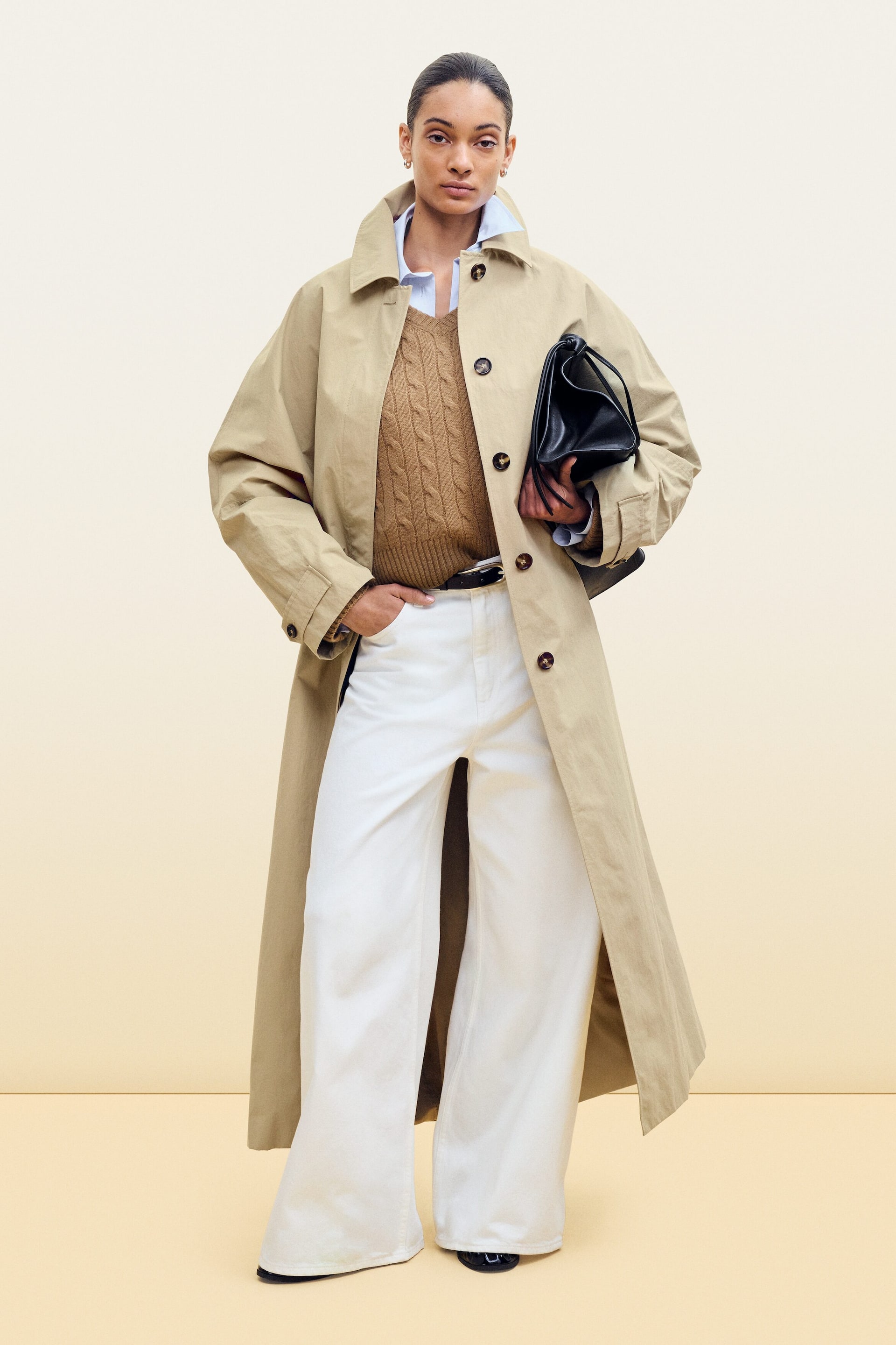 Grotere afbeelding bekijken: Een outfit bestaat uit een lange, lichtbruine trenchcoat die open wordt gedragen over een lichtblauw overhemd met kraag en een bruine kabelgebreide V-hals sweater vest. Witte wijde pijp broeken maken de onderste helft compleet, geaccentueerd met een donkere riem en een zwarte geplooide handtas.