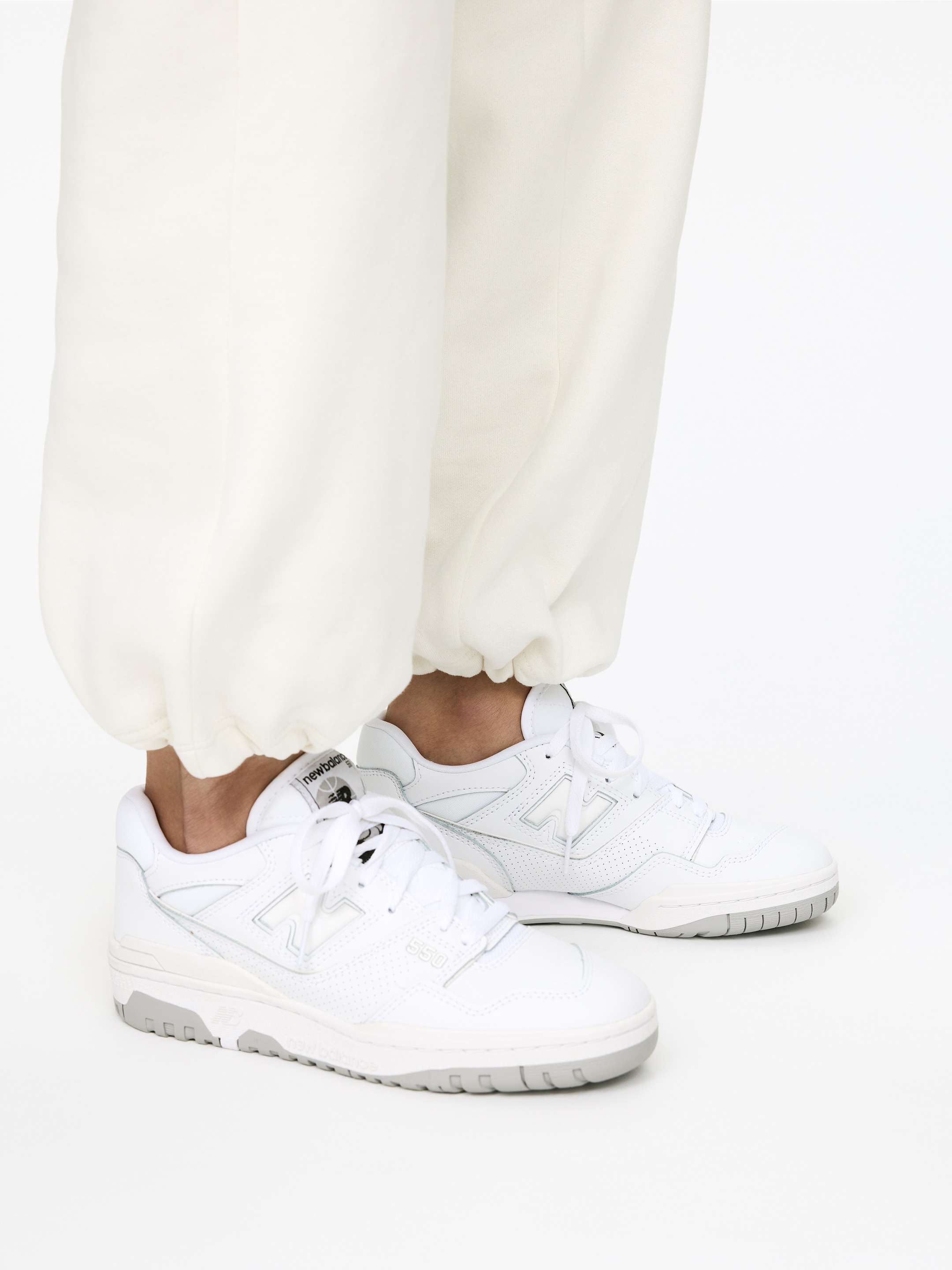 Agrandir l'image: Les baskets New Balance 550 blanches sont présentées sous un angle de côté avant, avec des tiges lisses et perforées blanches, un logo "N" blanc sur les côtés et un logo "new balance" sur la languette. Les lacets blancs sont passés à travers les œillets, et une semelle extérieure en caoutchouc gris offre un contraste. Un pantalon de survêtement de couleur crème est retroussé au-dessus de la cheville, ce qui cache partiellement le haut des baskets.