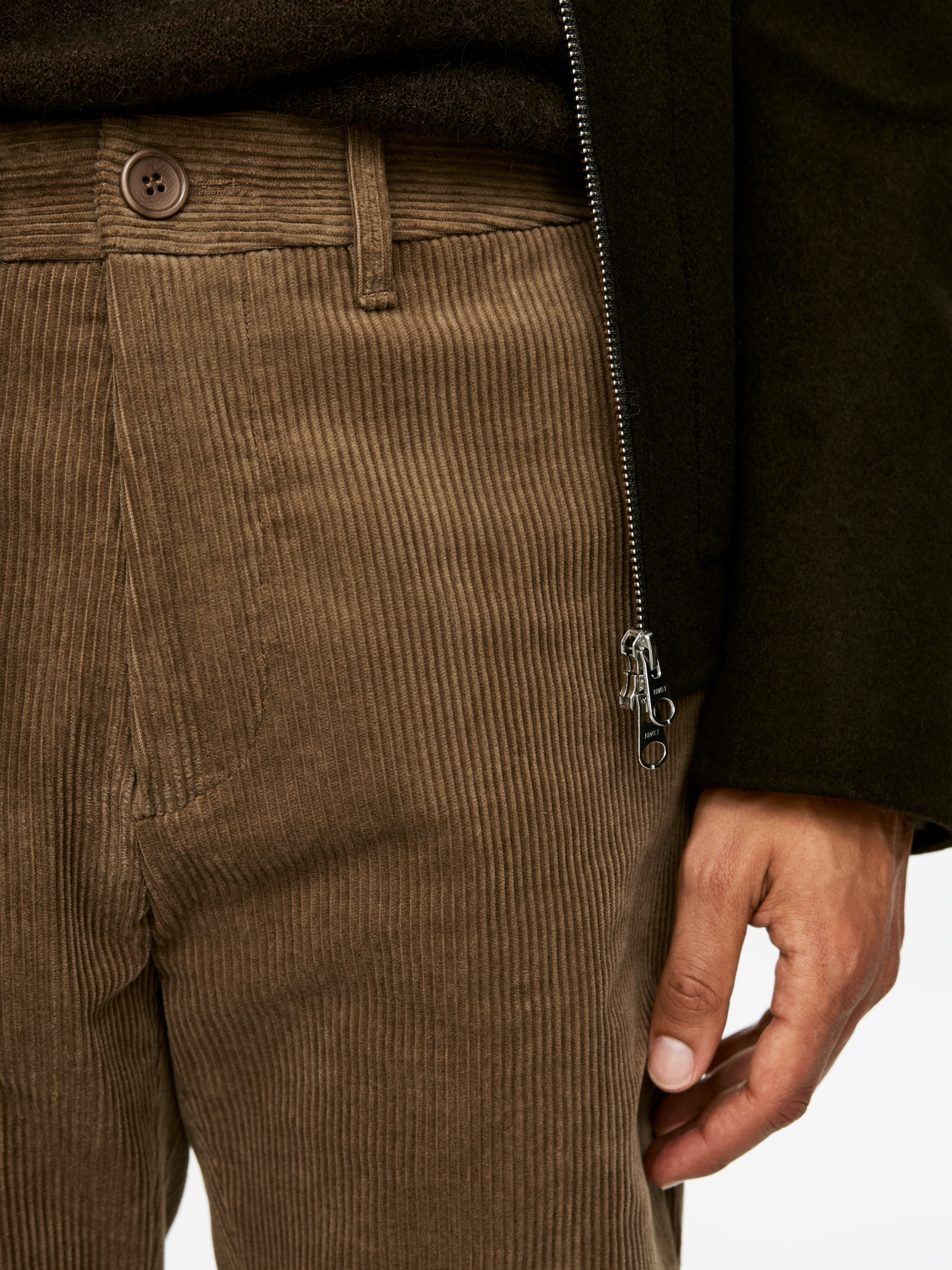 Visualizza immagine più grande: I pantaloni di velluto a coste marroni sono mostrati da un'angolazione all'altezza della parte superiore della coscia, con coste verticali prominenti, una chiusura a bottone marrone visibile e passanti per cintura in vita. Sono indossati, stratificati con un top scuro a maglia e una giacca scura con cerniera, con una mano che riposa vicino a una tasca laterale, evidenziando la vestibilità e la texture.