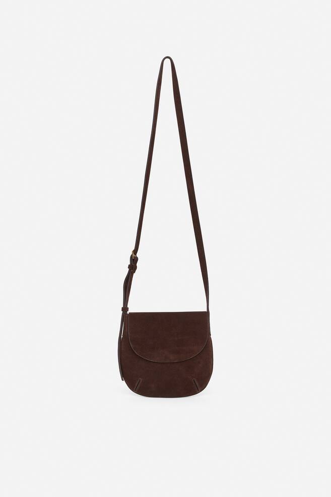 Se muestra un bolso bandolera de ante marrón oscuro de frente, con una solapa redondeada y un cuerpo principal curvado. Se aprecia una costura sutil en forma de dardo en la parte inferior delantera, y una correa delgada y ajustable con una pequeña hebilla se extiende desde la parte superior. - 1