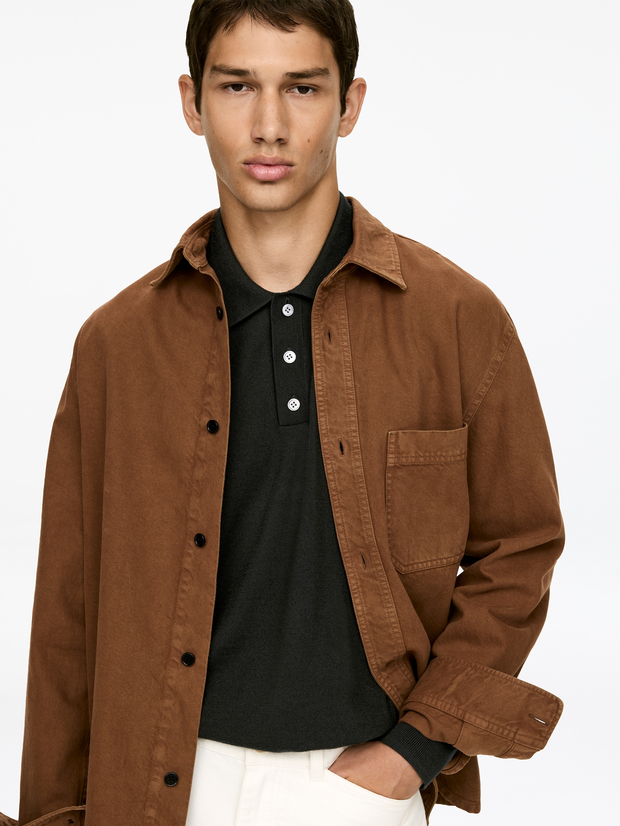 Grösseres Bild ansehen: Ein braunes Overshirt ist von vorne zu sehen und offen über ein dunkles Strick-Poloshirt gestylt. Es verfügt über einen spitzen Kragen, eine Knopfleiste mit dunklen Knöpfen und eine aufgesetzte Tasche, wobei die langen Ärmel hochgekrempelt sind, um die gerippten Bündchen des Unterhemdes zu zeigen.